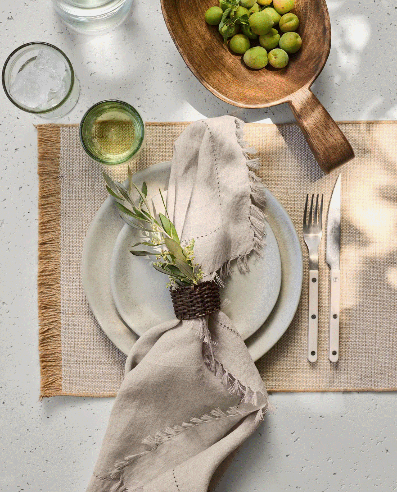 Table setting with a white ceramic plate, beige napkin with greenery, fork and knife with white polka dot handles, a glass of white wine, two glasses of water, a wooden bowl of green grapes, and a glass of water on a woven placemat.