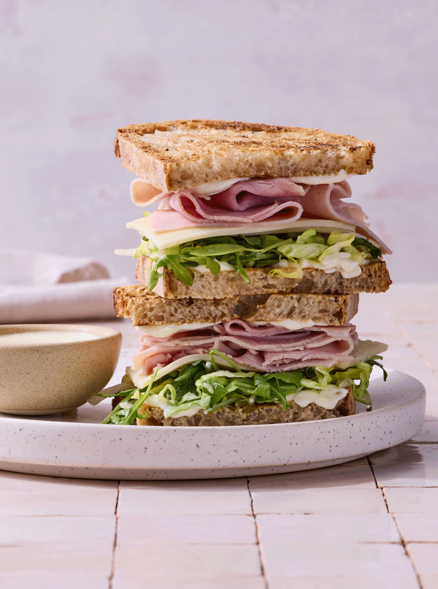Stacked deli sandwiches with lettuce, turkey, ham, cheese, and toasted bread on a white plate, with a small bowl of dressing or sauce nearby.