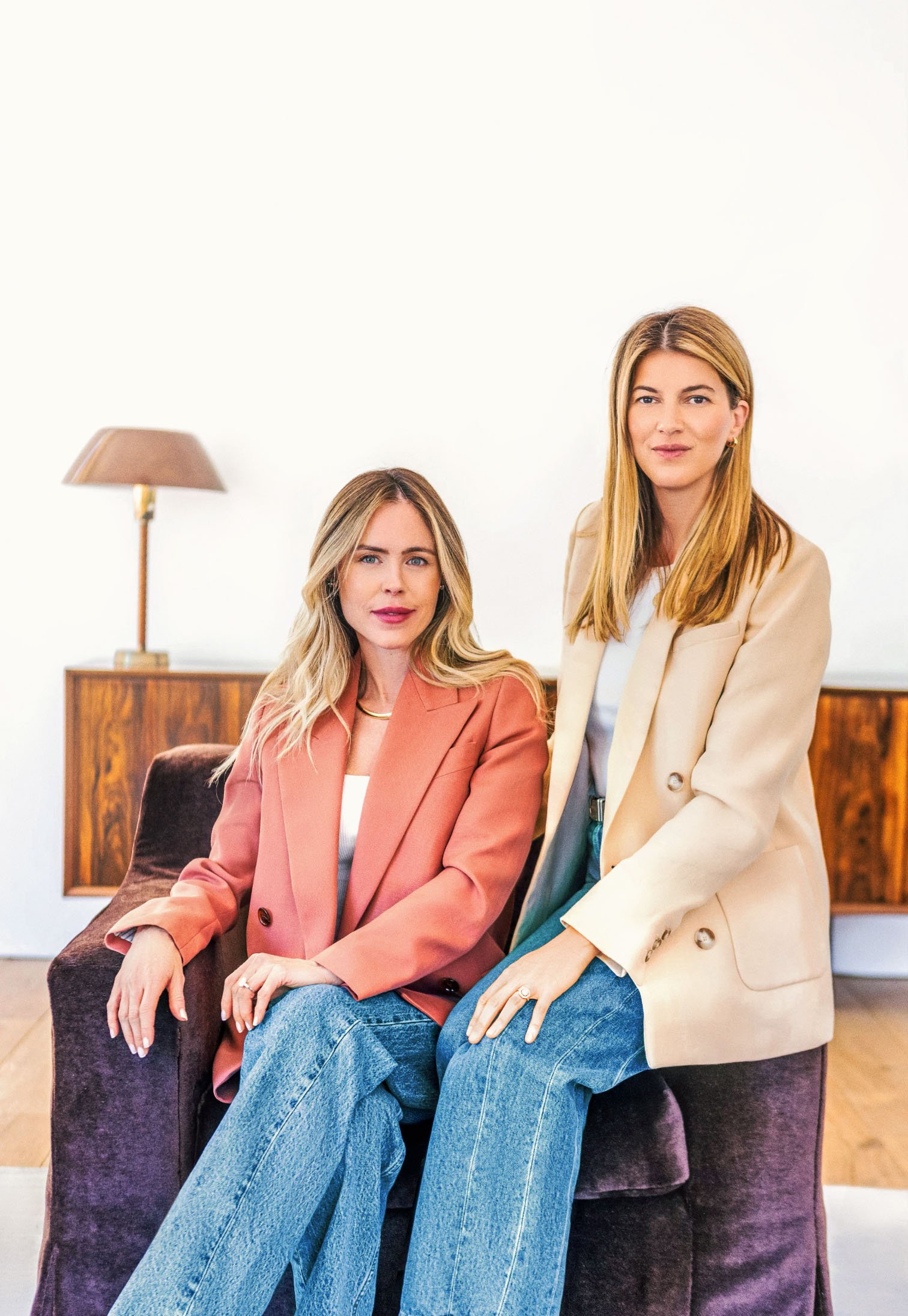 Two women with blonde hair wearing blazers and jeans sitting on a purple velvet armchair in a room with a wooden sideboard and lamp in the background.