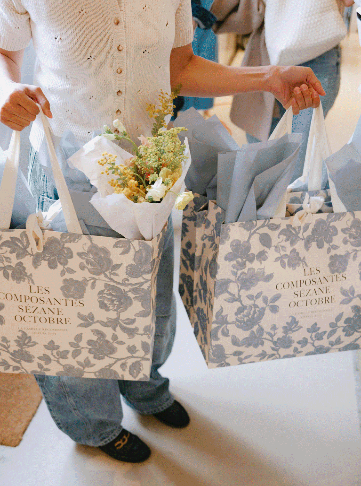 Two people holding floral shopping bags with flowers and gray tissue paper inside at a store.