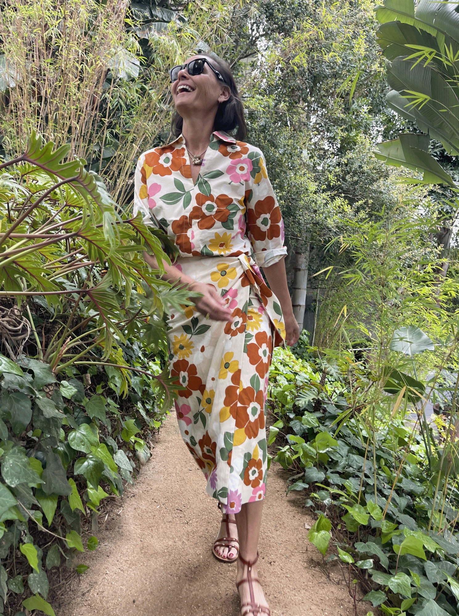 An older woman walking through a lush garden, smiling and enjoying the outdoors, wearing a colorful floral dress and sunglasses.