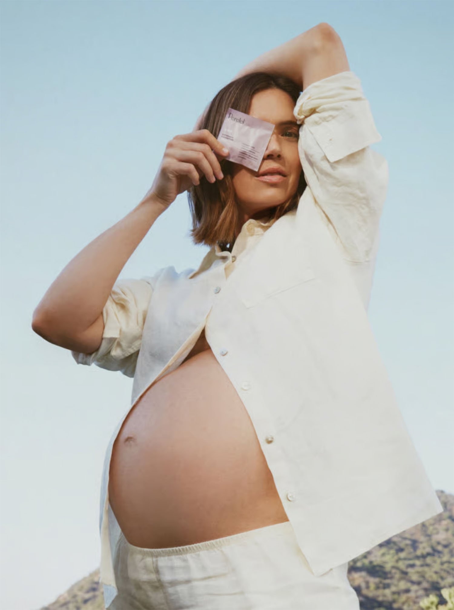 A pregnant Mandy Moore woman holding a plane ticket and posing outdoors against a clear sky.