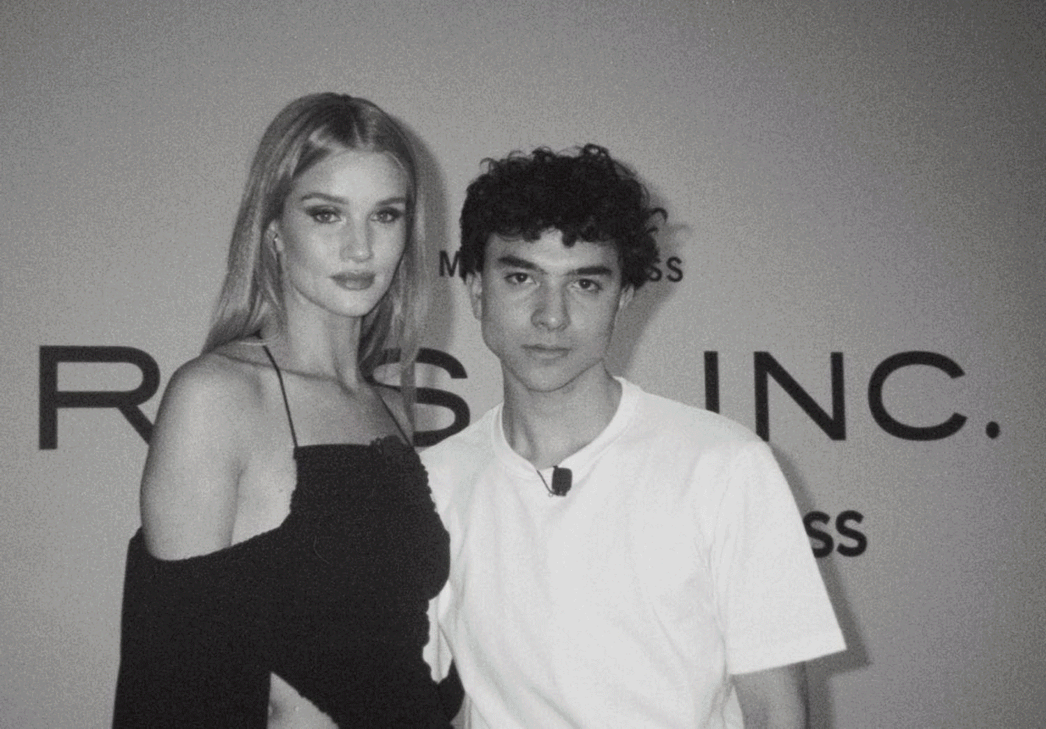 A black and white photo of a woman with long hair wearing a dark off-shoulder dress and a young man with curly hair wearing a white T-shirt, standing together in front of a backdrop with text.