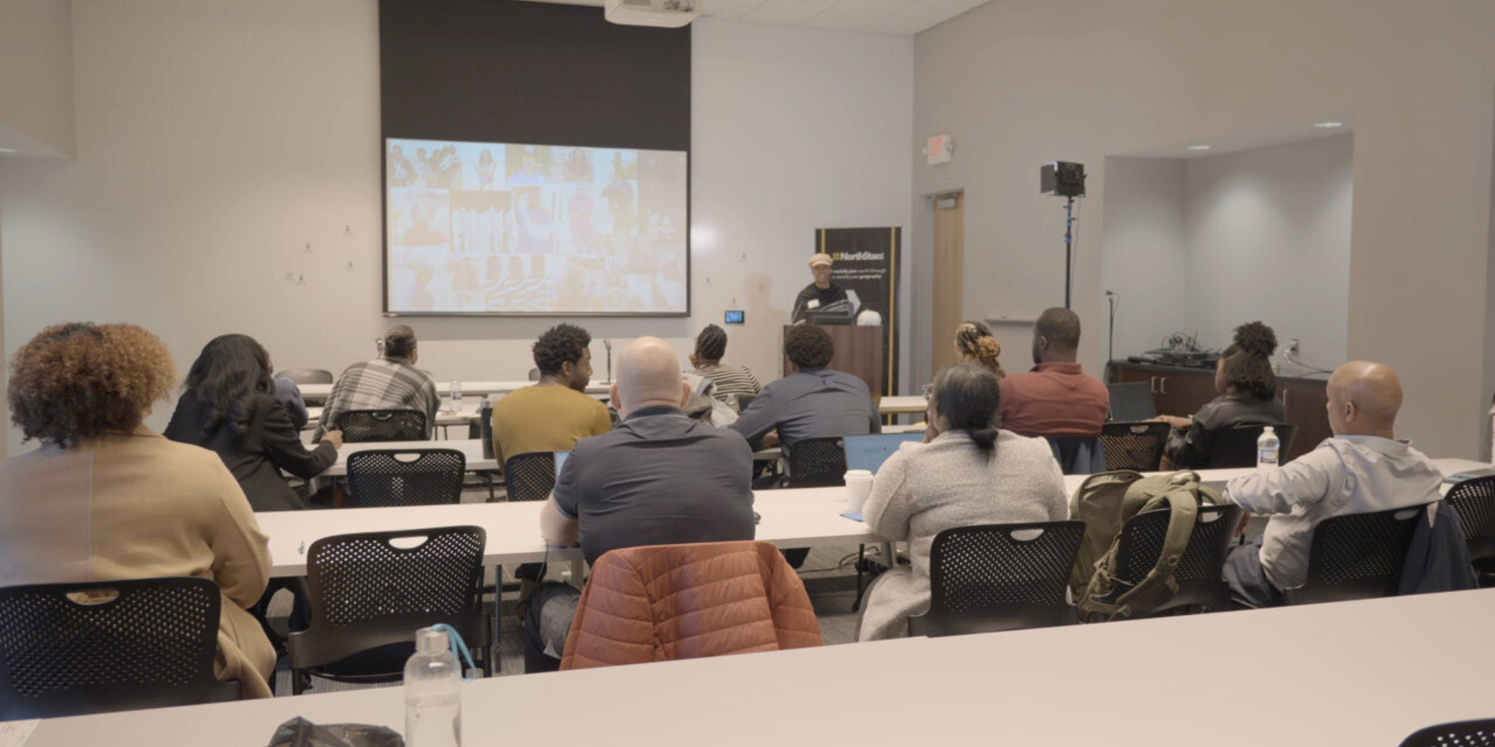 People attending a presentation or lecture in a classroom or conference room, facing a speaker at the front with a large screen behind them.