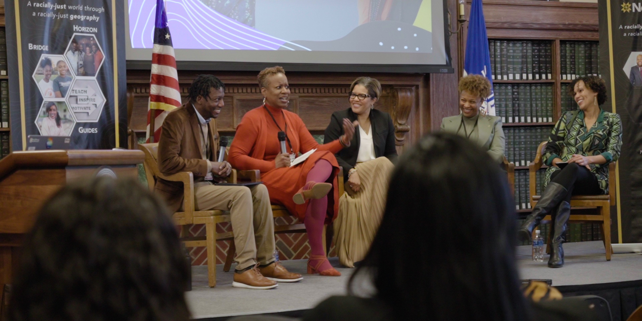 Panel discussion with five women seated on a stage, holding microphones. The background features flags, bookshelves, and a large screen displaying graphics. Audience members are in the foreground.