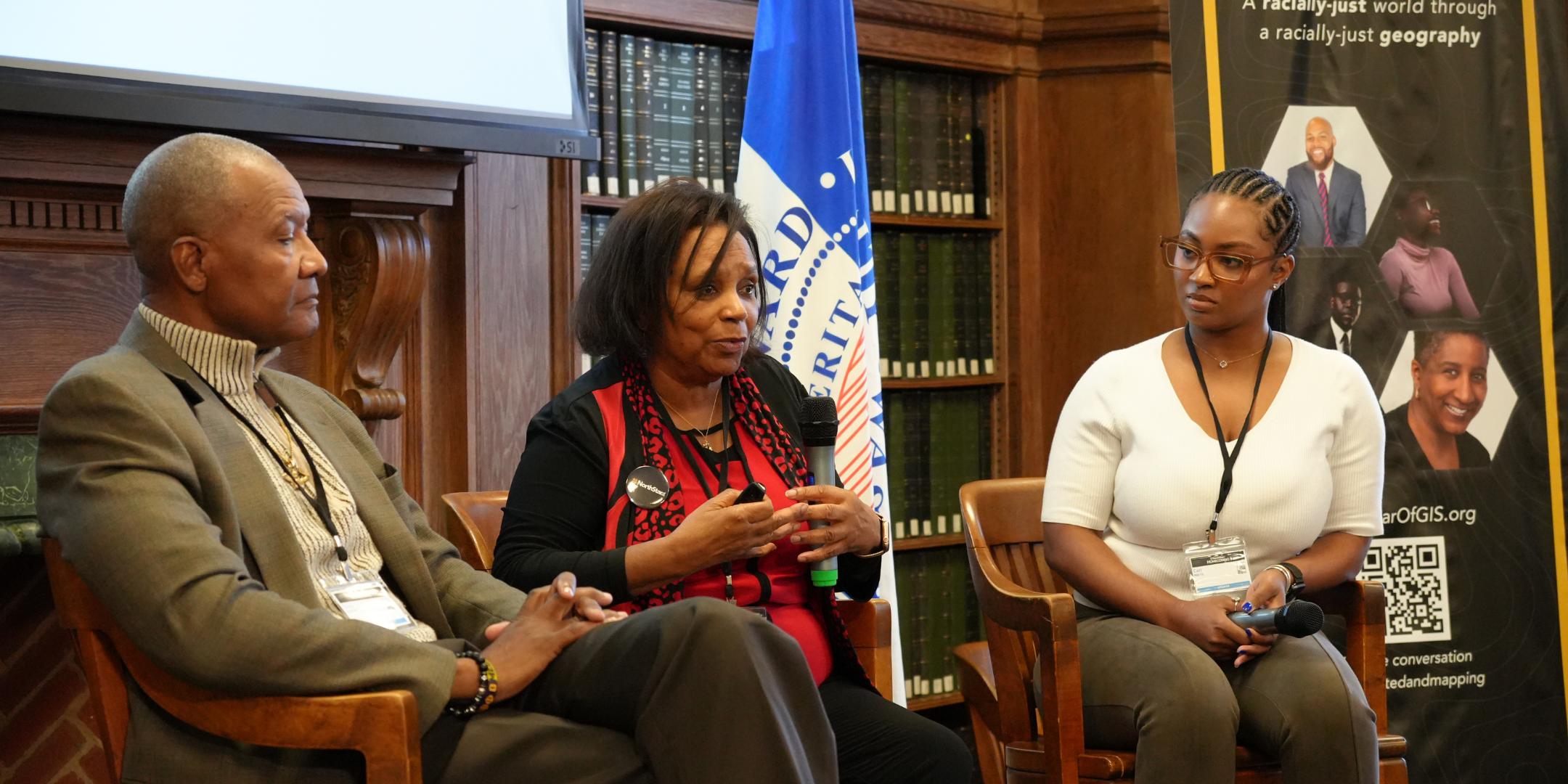 Three women seated on wooden chairs in a panel discussion. The woman in the middle is speaking into a microphone, wearing a black and red outfit. The woman on the left is dressed in a brown blazer with a striped turtleneck, while the woman on the rig