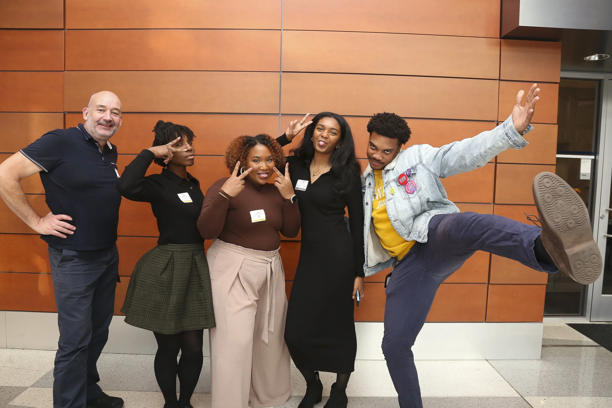 Group of five diverse people posing together indoors against a wooden paneled wall, making peace and playful gestures.