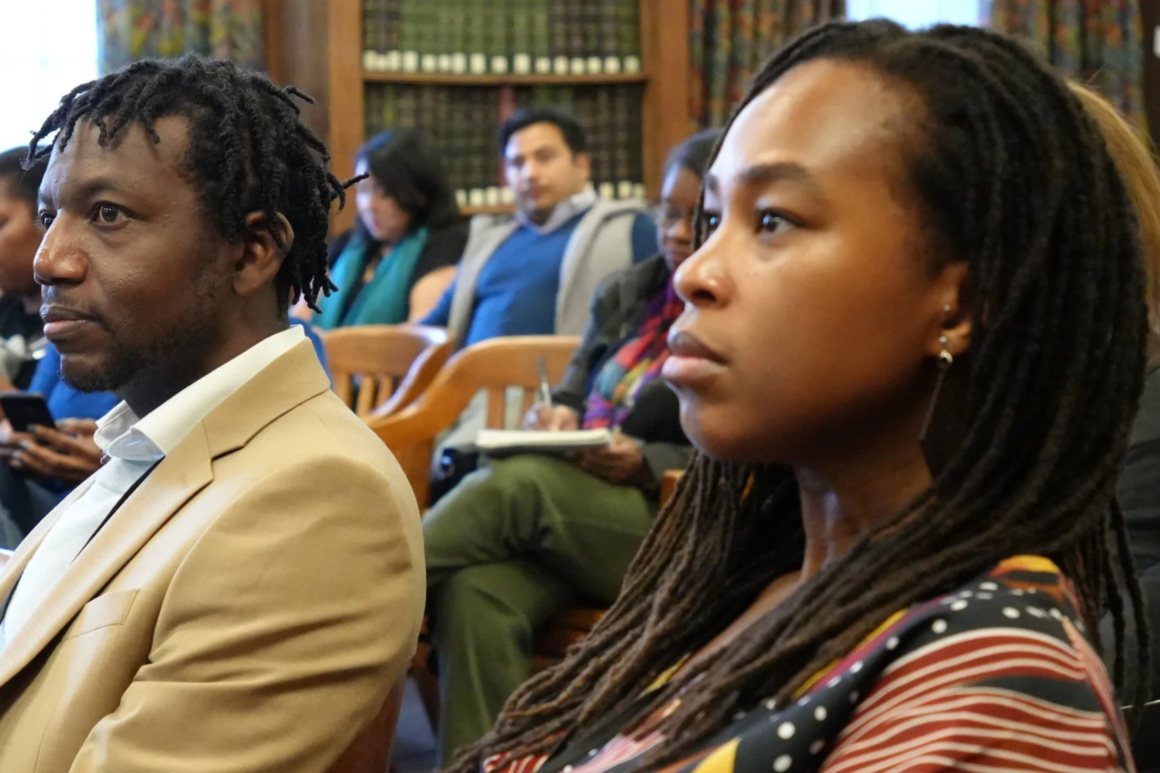 People attending a formal event or seminar, sitting in a room with wood-paneled bookshelves, listening attentively.