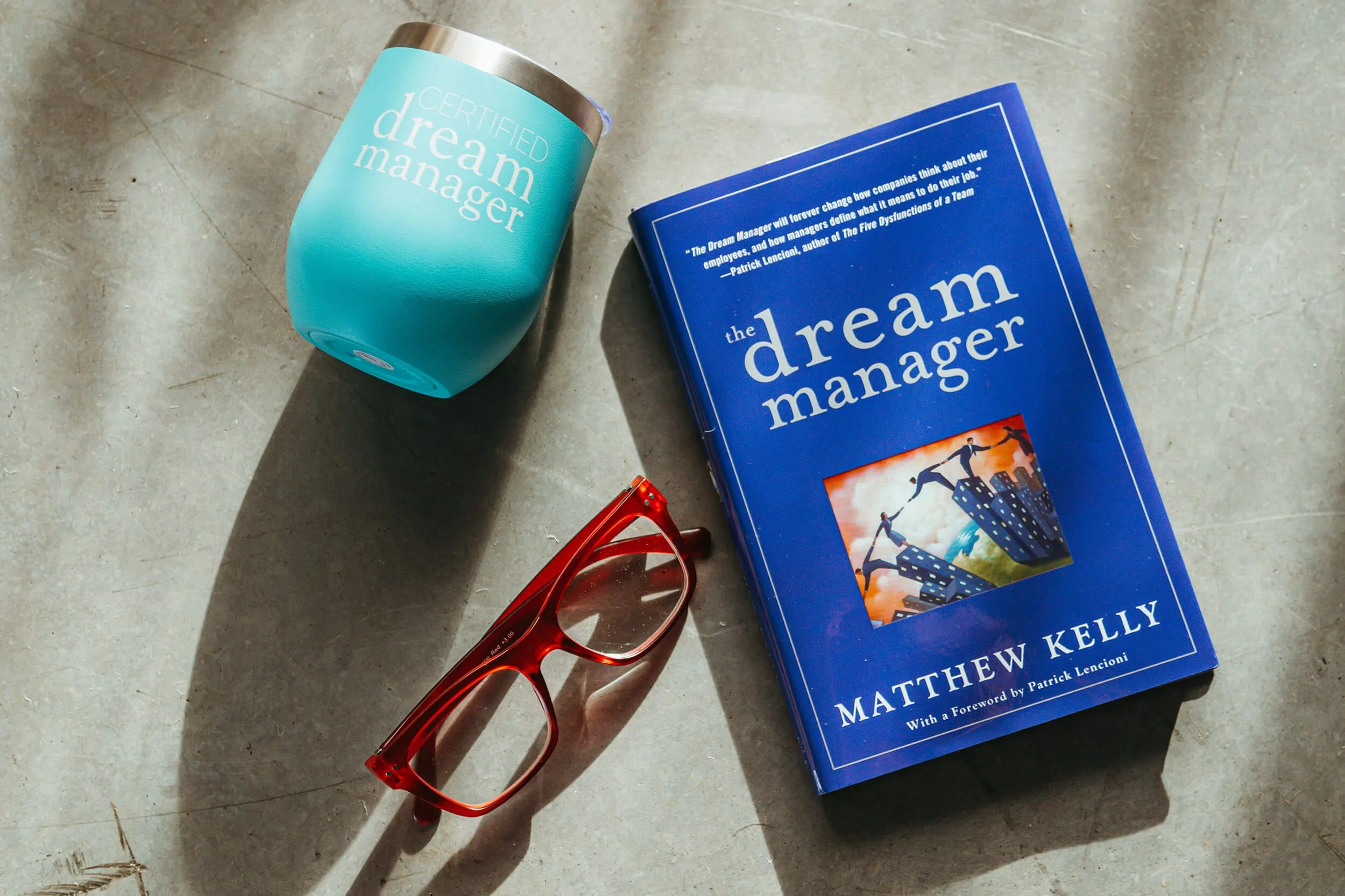 A blue book titled "The Dream Manager" by Matthew Kelly, a blue tumbler with a lid labeled "Certified Dream Manager," and a pair of red glasses on a concrete surface, with sunlight casting shadows.