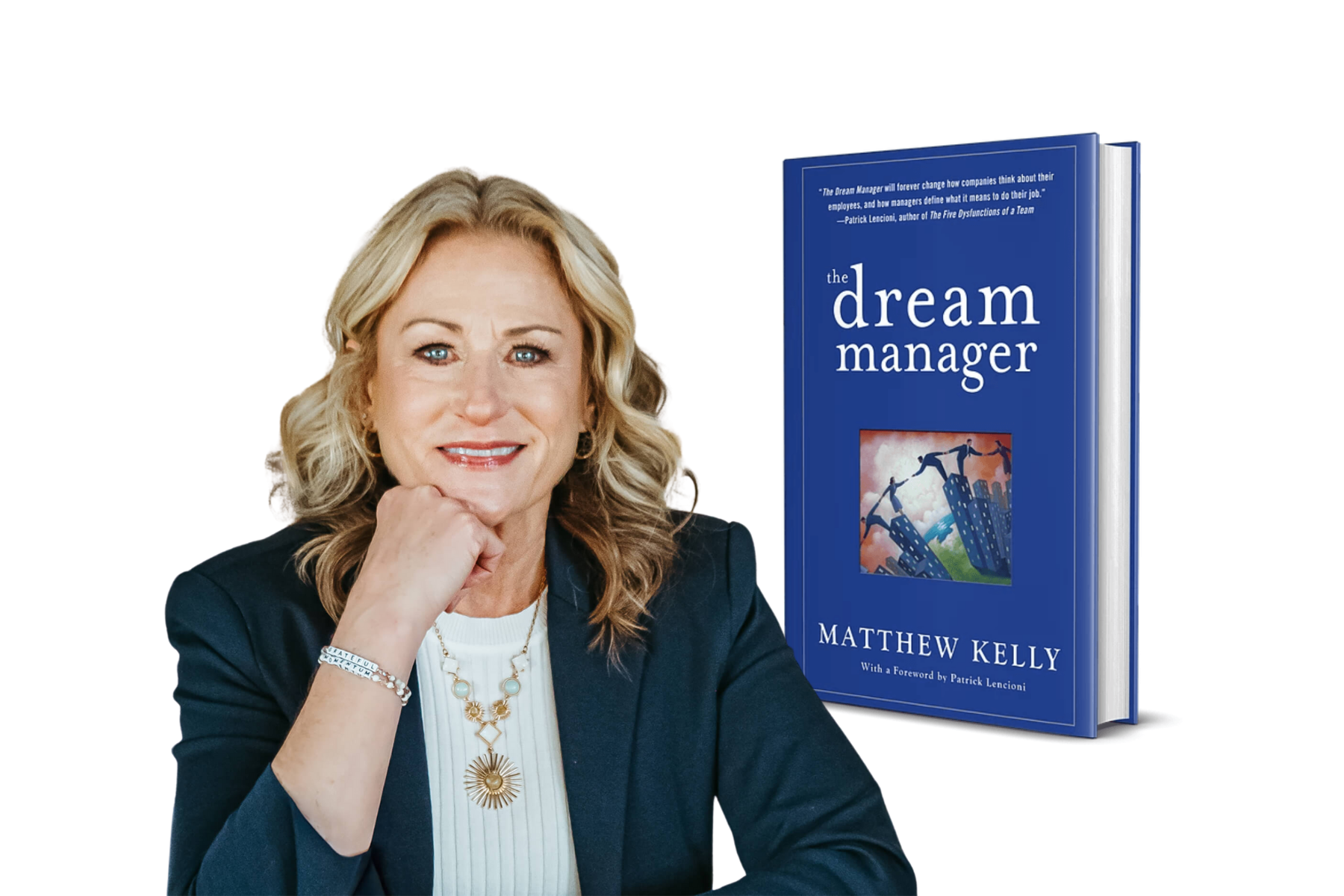 A woman with blonde hair smiling, resting her chin on her hand, wearing a black blazer, white top, and jewelry, beside a blue book titled 'The Dream Manager' by Matthew Kelly.