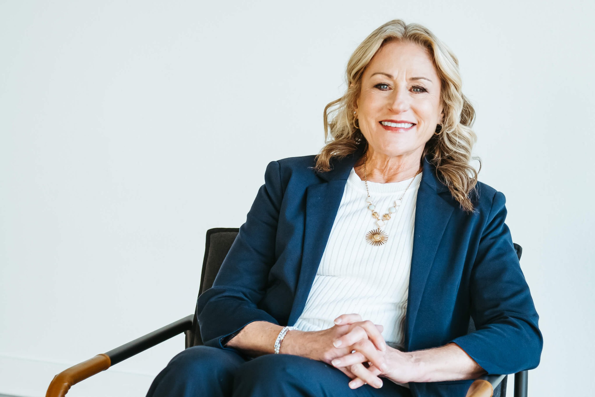A smiling woman with blonde hair dressed in a navy blue blazer and white blouse, sitting in a black chair with wooden armrests against a plain white background.