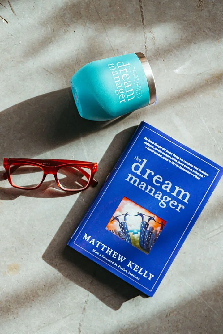 A blue insulated tumbler with the words 'certified dream matcher,' a pair of red glasses, and a blue book titled 'the dream manager' by Matthew Kelly are arranged on a light-colored surface.