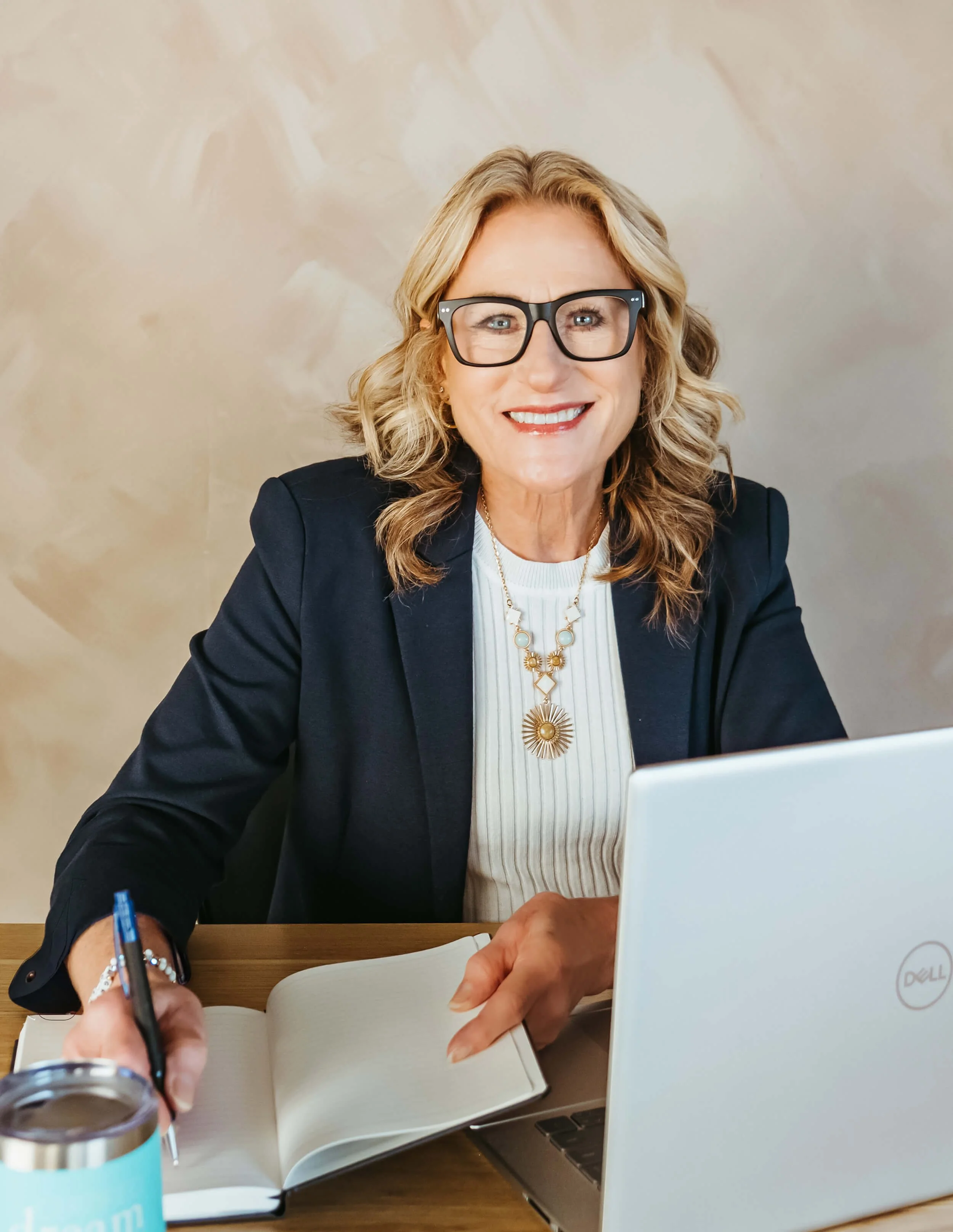Smiling professional woman with glasses, wearing a dark blazer and a white top, sitting at a desk with a notebook, pen, and a laptop.