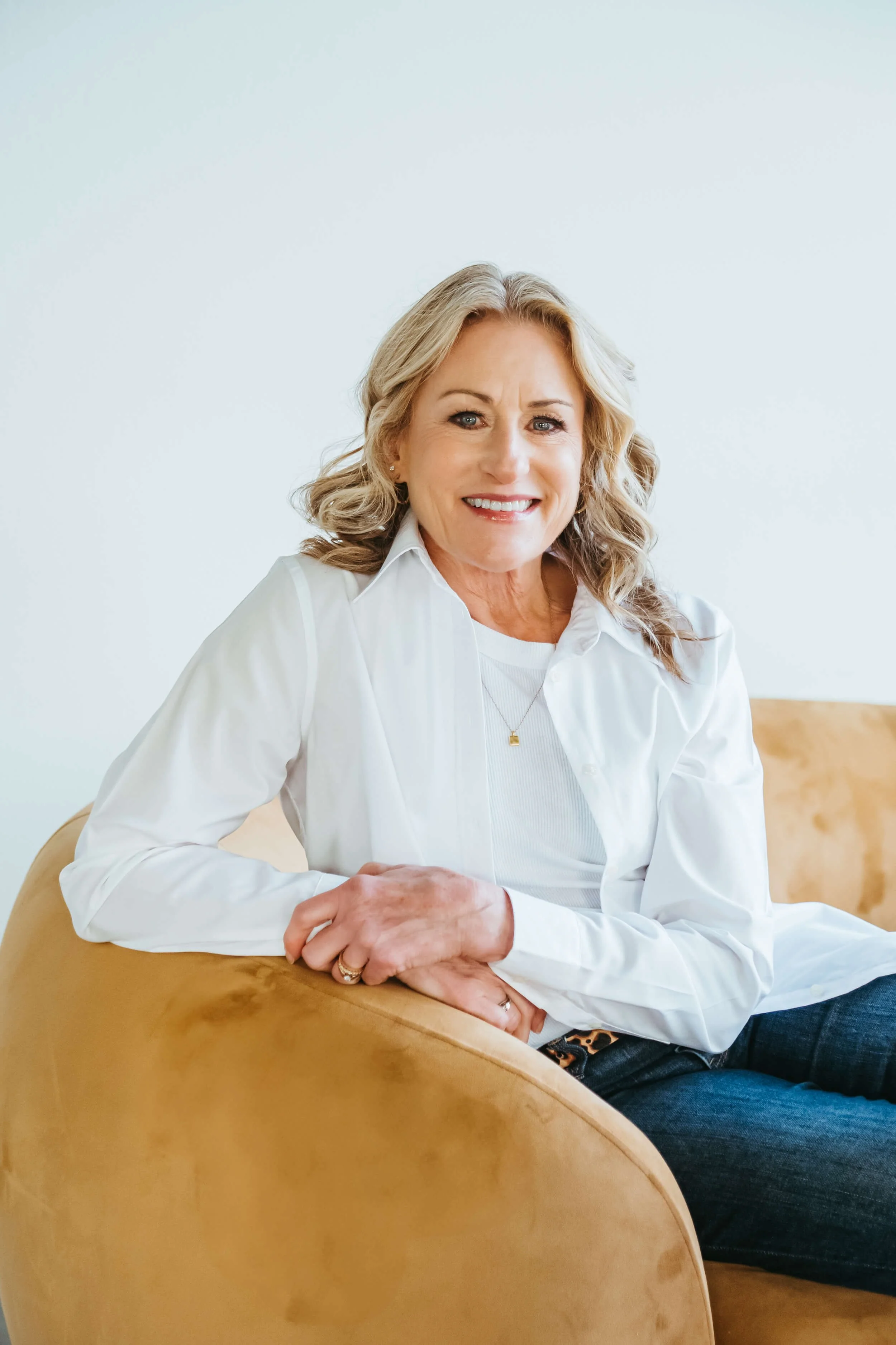 A woman with blonde curly hair sitting on a tan sofa, smiling at the camera, wearing a white shirt and dark jeans, in a bright room with a white background.