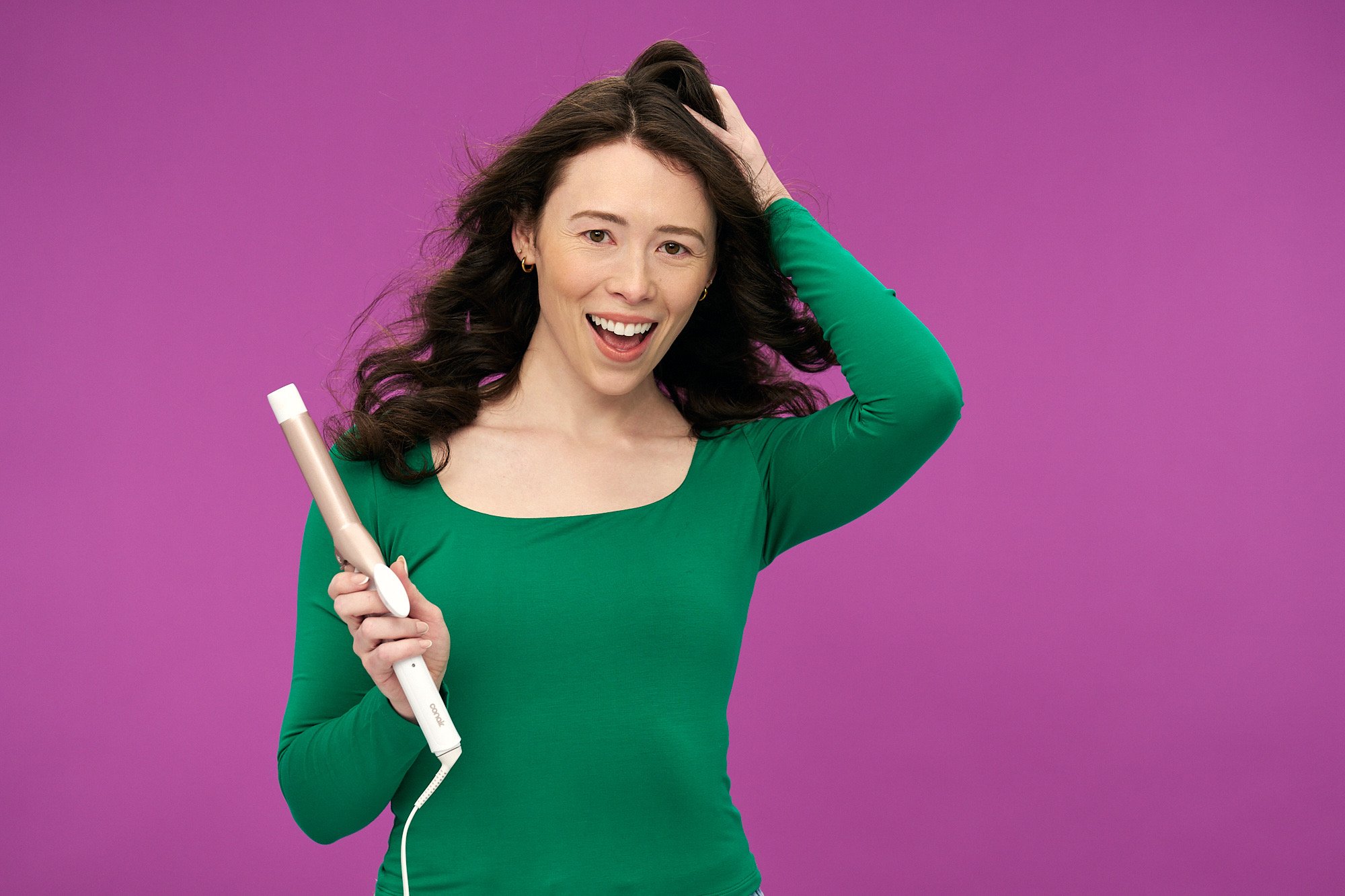 purple background with brunetter girl in green long sleeve tshirt holding pink and white conair curling iron