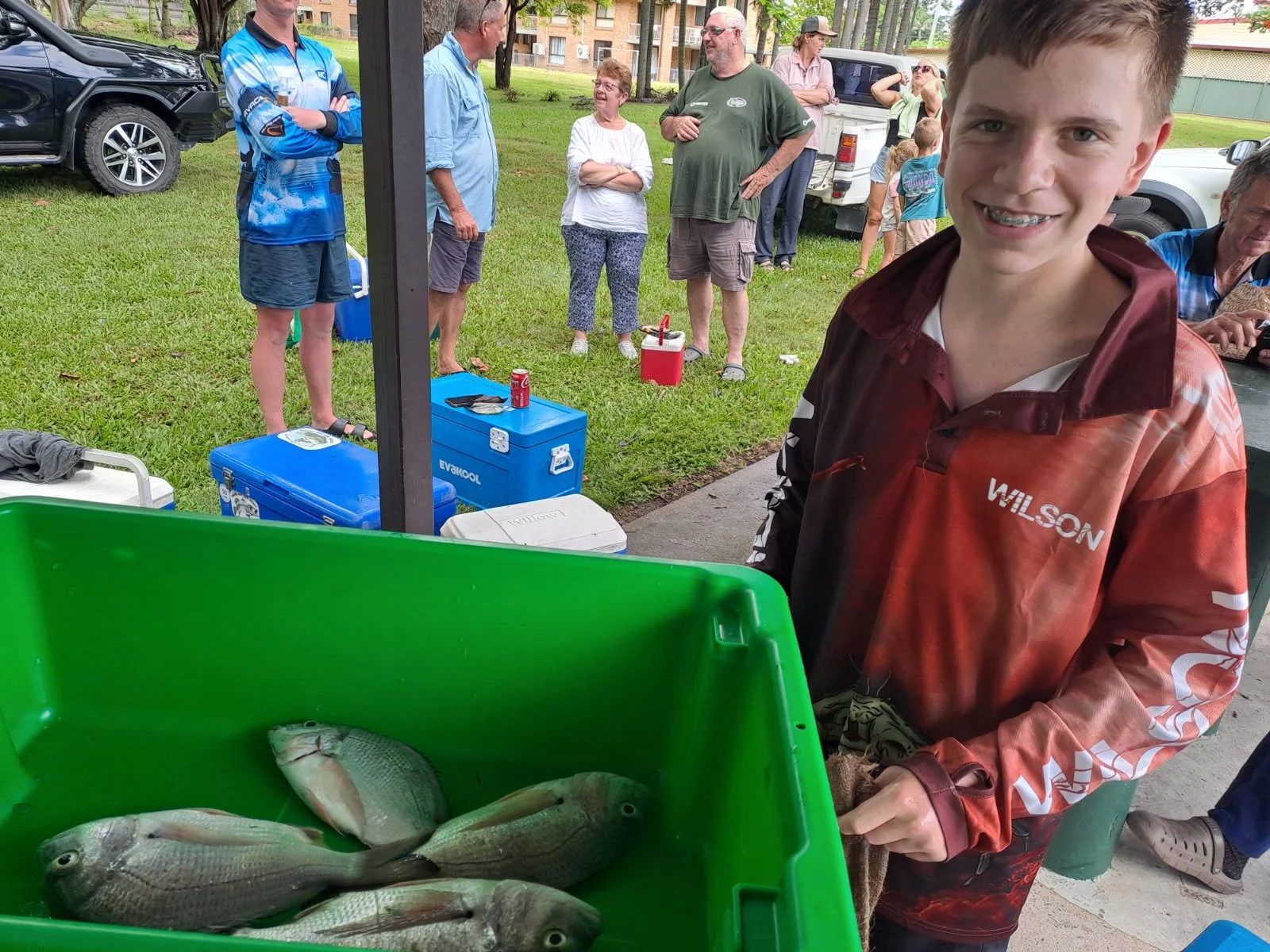 A young boy with braces smiling at a fishing event with a green container of fish, and people in the background near parked cars and coolers.