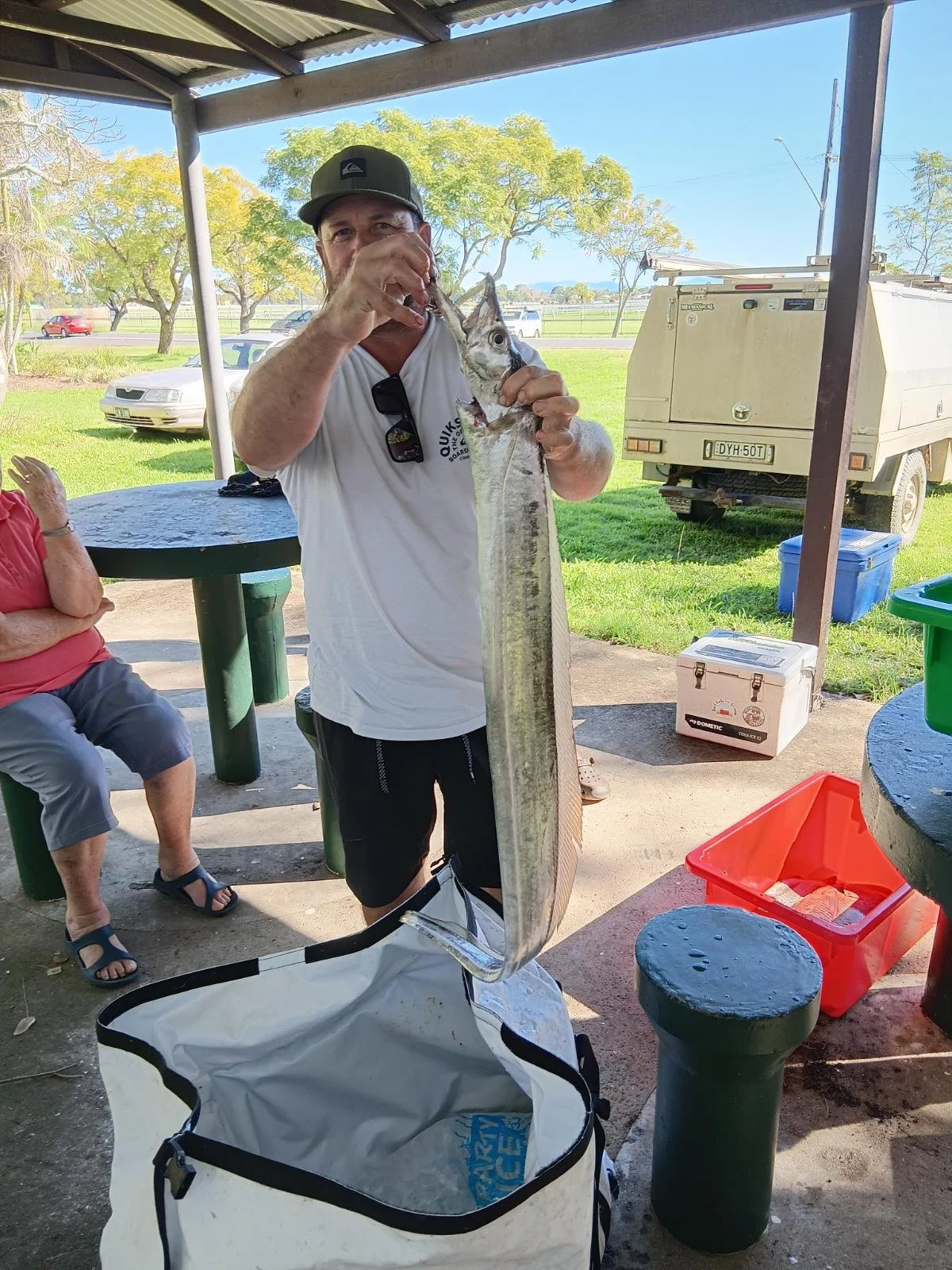A man holding a large fish with a woman sitting nearby at an outdoor covered picnic area.