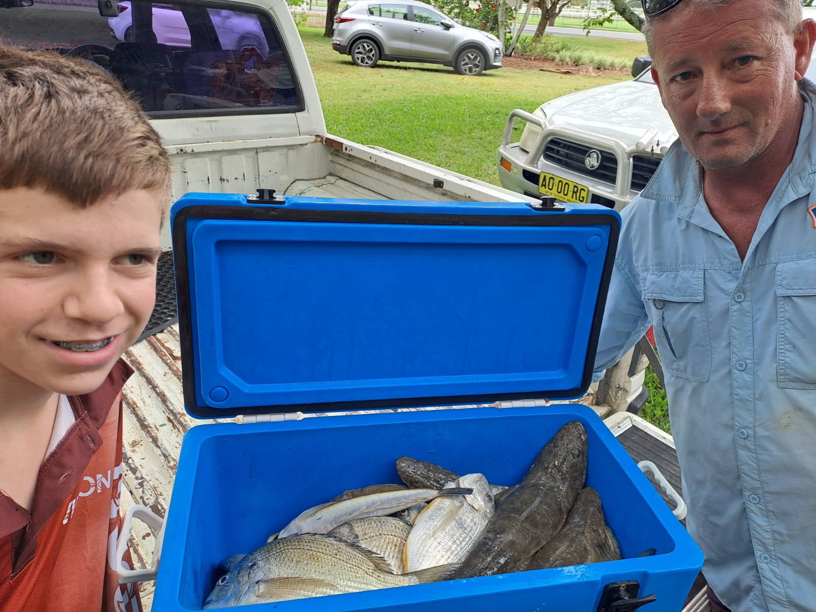 A boy and a man standing next to a blue cooler filled with freshly caught fish in the back of a white pickup truck.