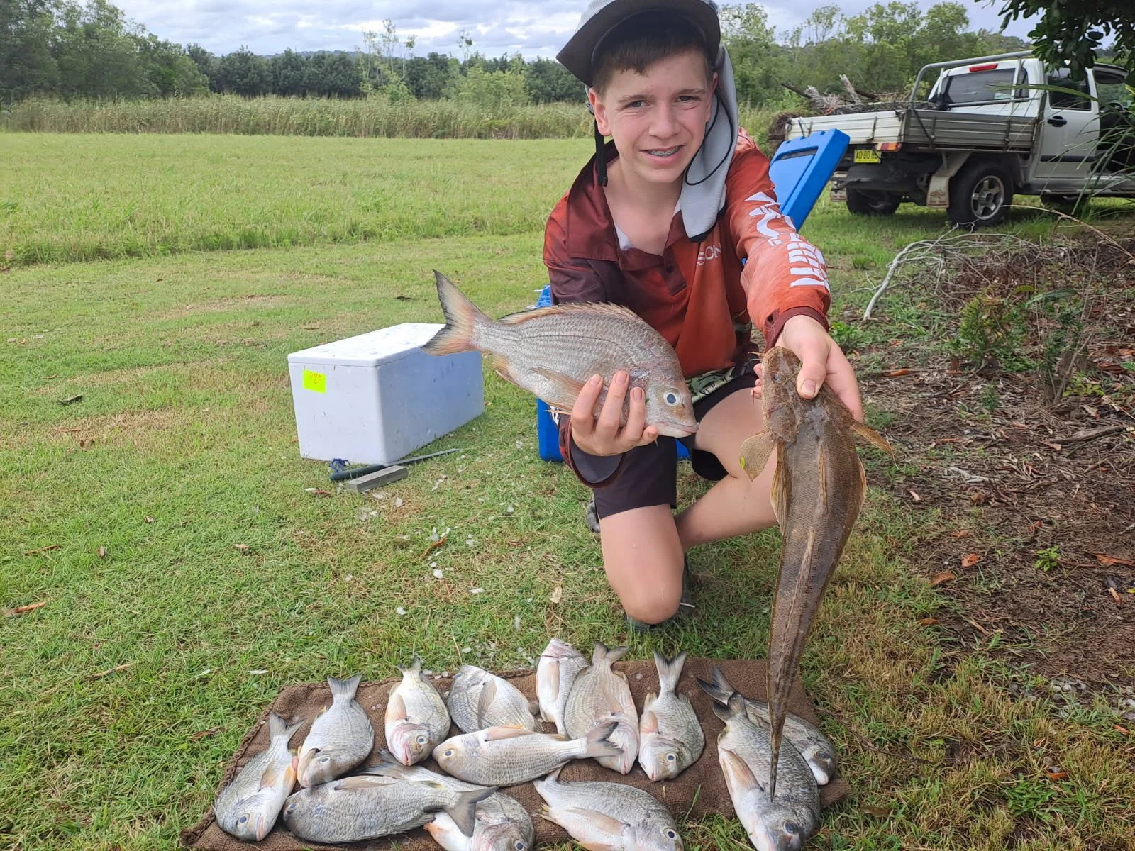 A young boy kneeling on grass, holding a large flathead fish in one hand and a red drum fish in the other, surrounded by a collection of other fish on a mat. He is outdoors in a field with trees and a pickup truck in the background.