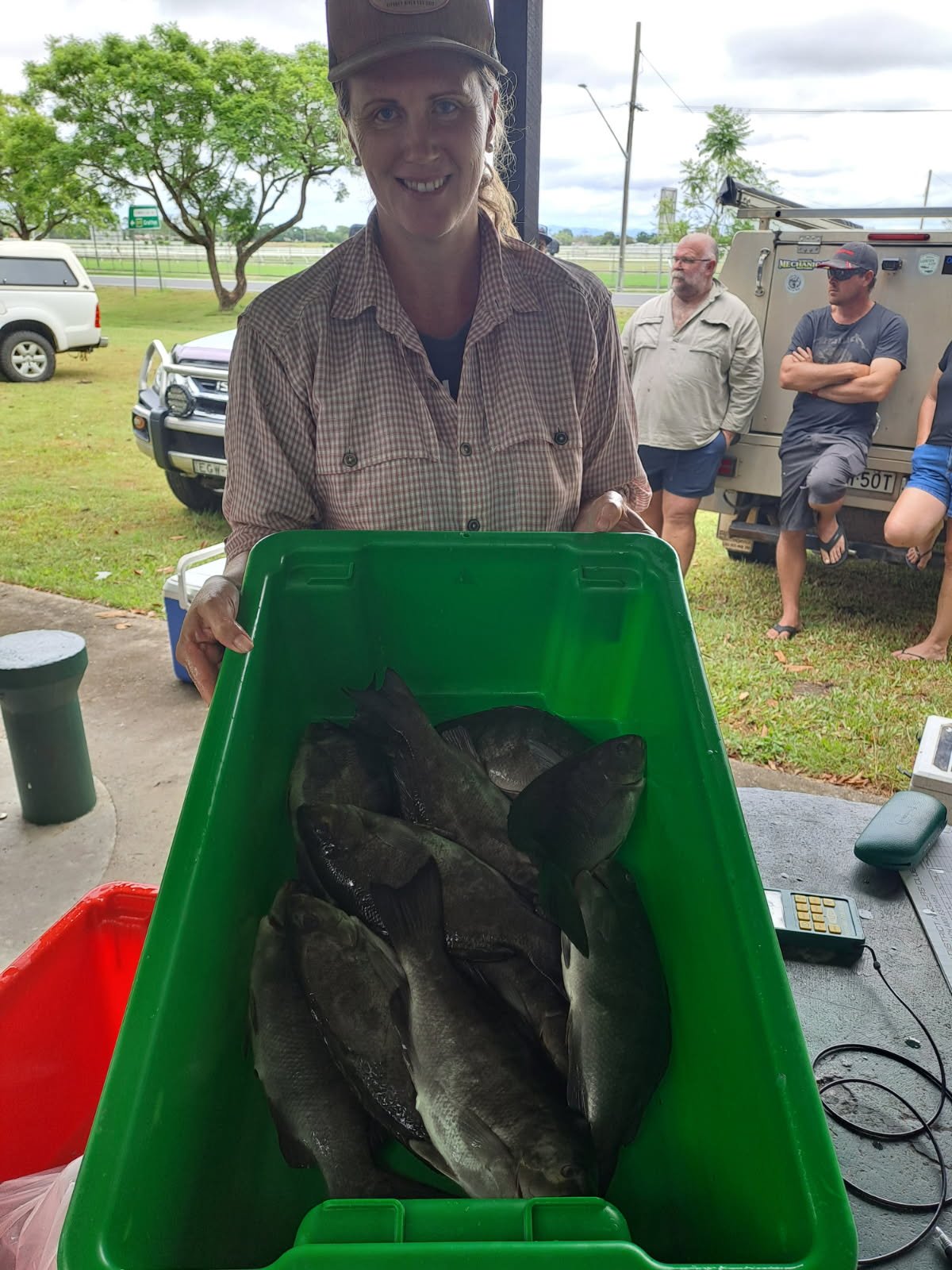 A woman with a baseball cap holding a green container filled with fish, smiling at the camera. In the background, there are three men and various vehicles outdoors near a grassy area with trees and cloudy sky.