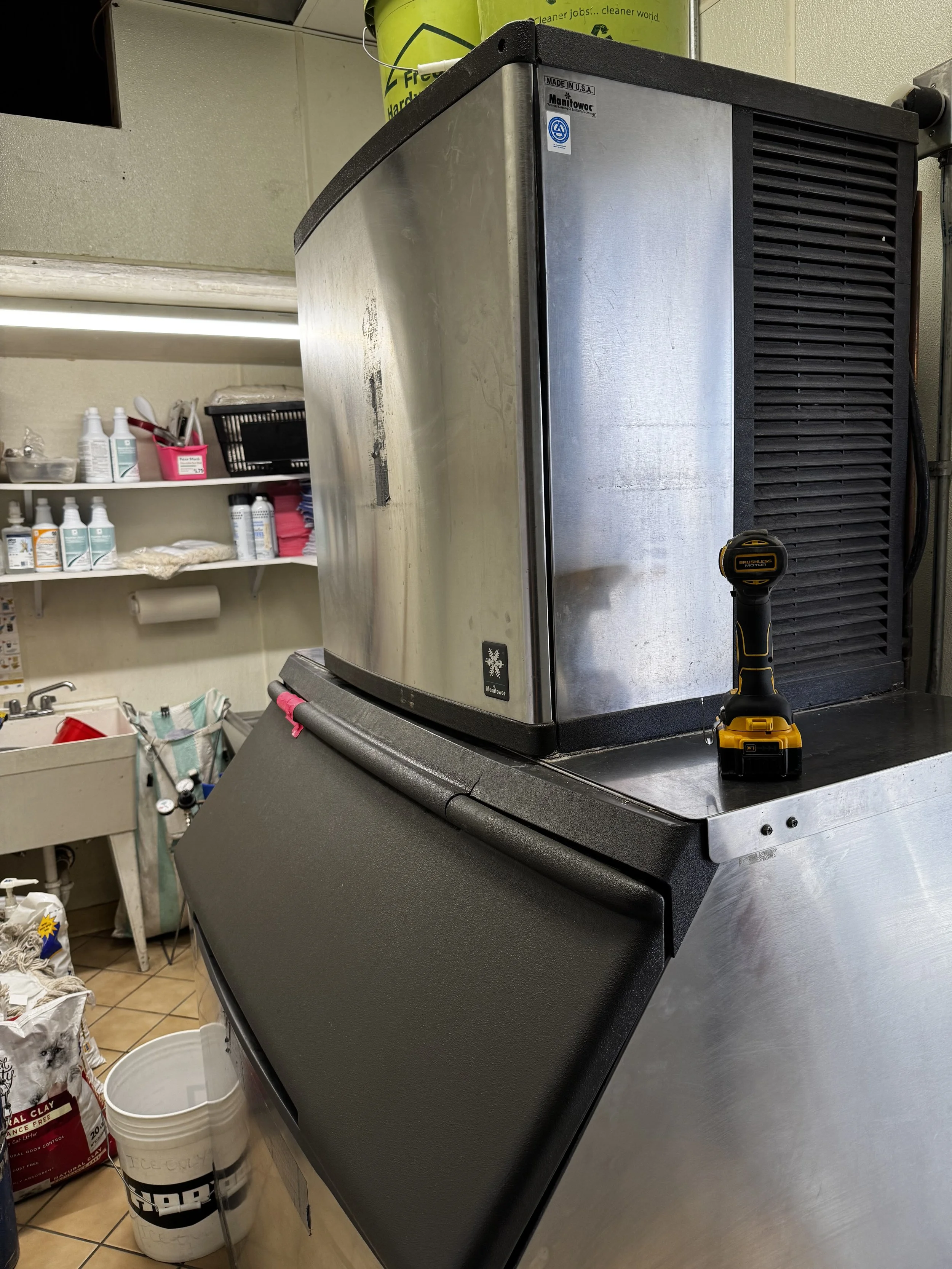 Commercial freezer with a cordless drill on top, located in a cleaning or maintenance room with shelves holding cleaning supplies and storage containers.