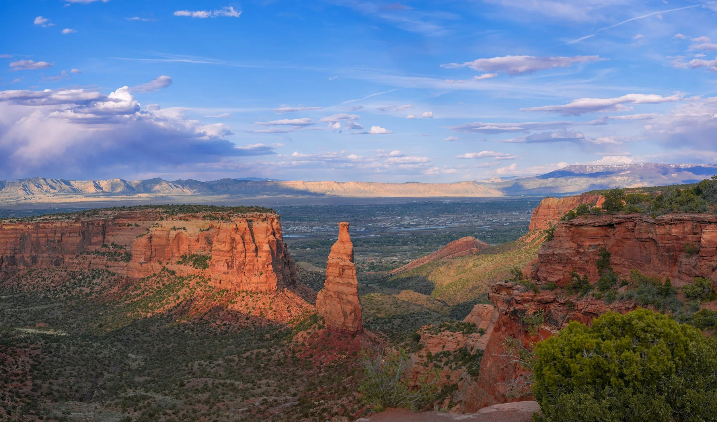 Red rock formations, including a prominent spire, in a canyon with green vegetation, under a partly cloudy sky.