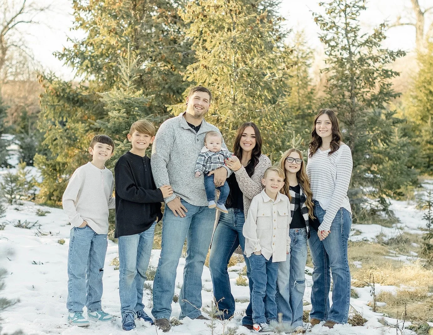 Family of nine standing outdoors on snow-covered ground with evergreen trees in the background, smiling at the camera.