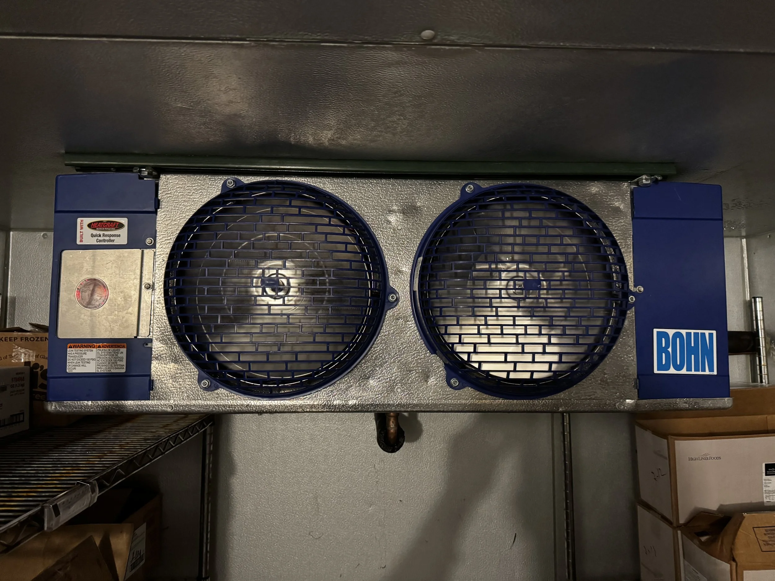 Metal refrigeration unit with two circular blue fans on top, labeled 'BOHN,' with control panel on the left and stored boxes below.