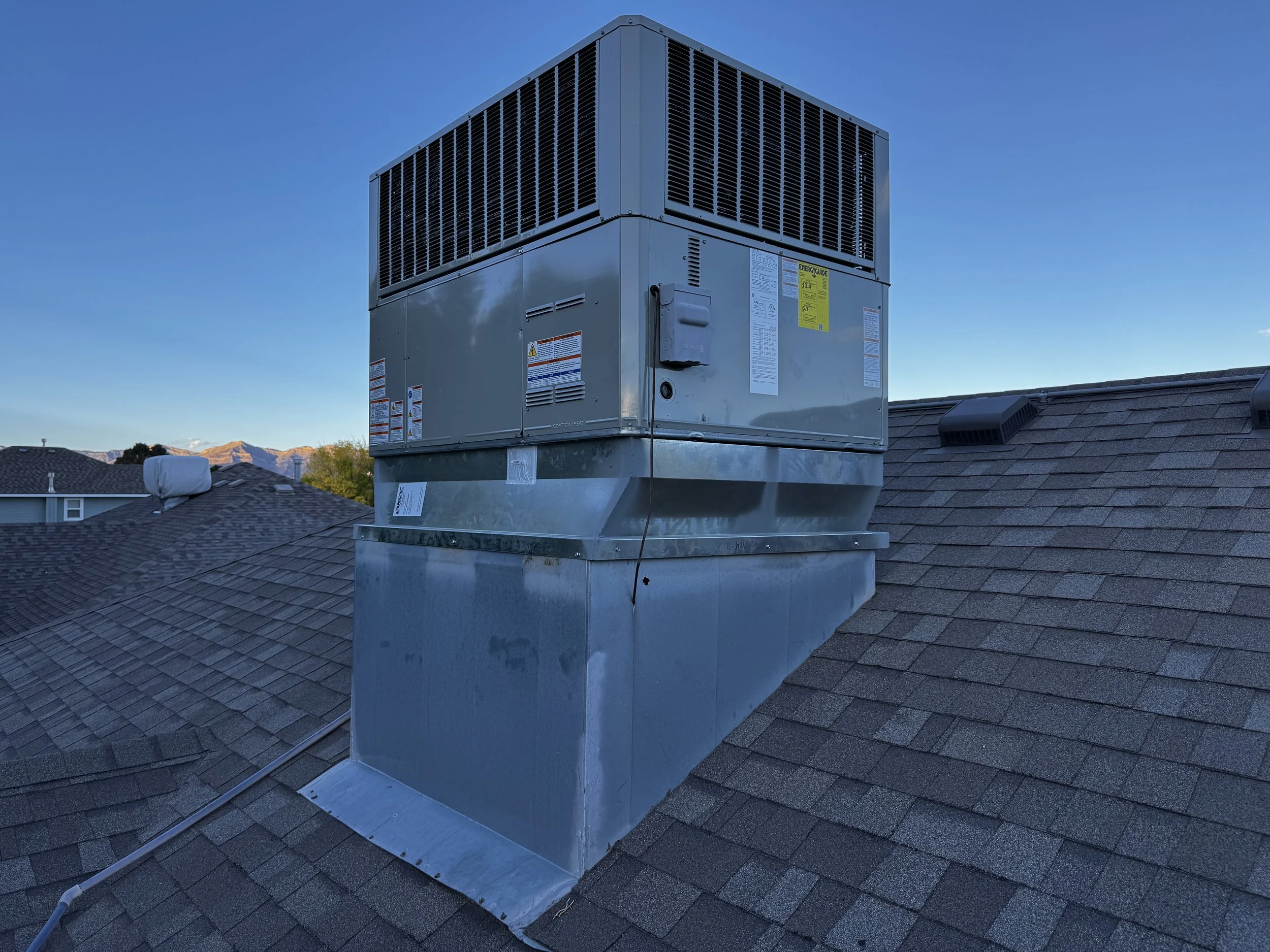 A rooftop HVAC unit installed on a shingled roof under a clear blue sky with distant mountains.