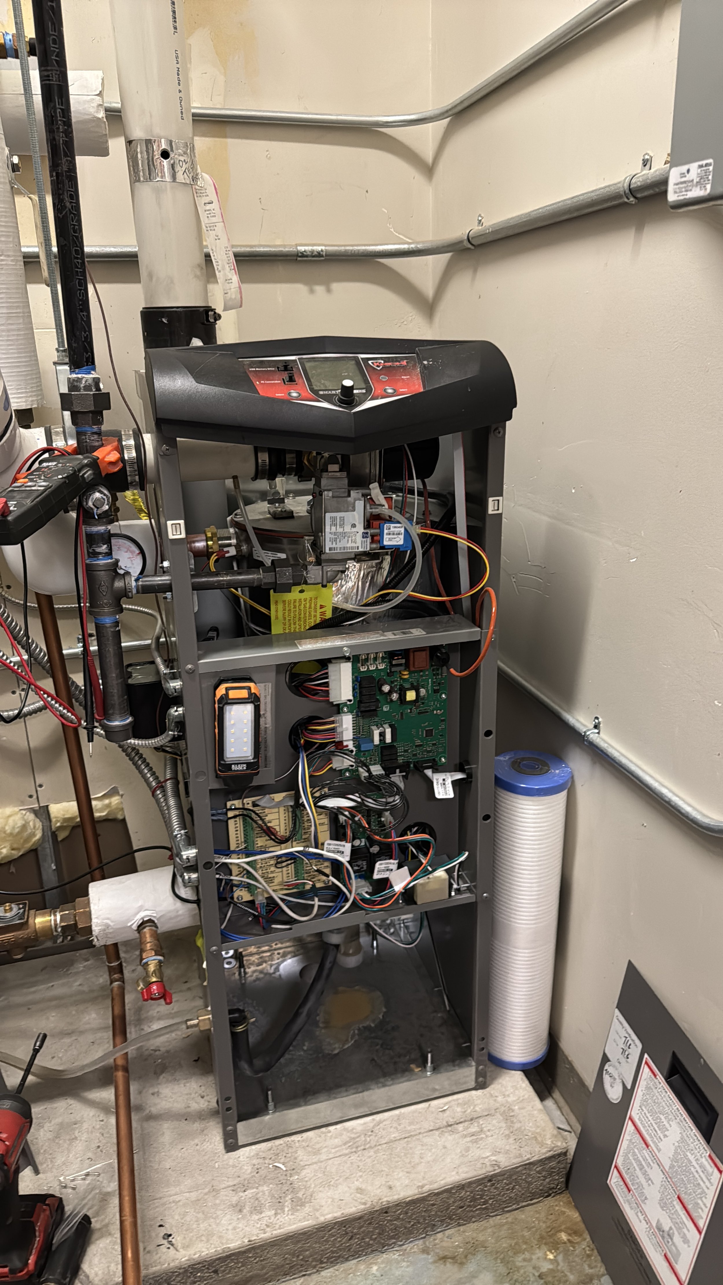 Open electronic panel revealing internal circuit boards, wires, and components, with pipes and a filter on the right side, in a utility room.