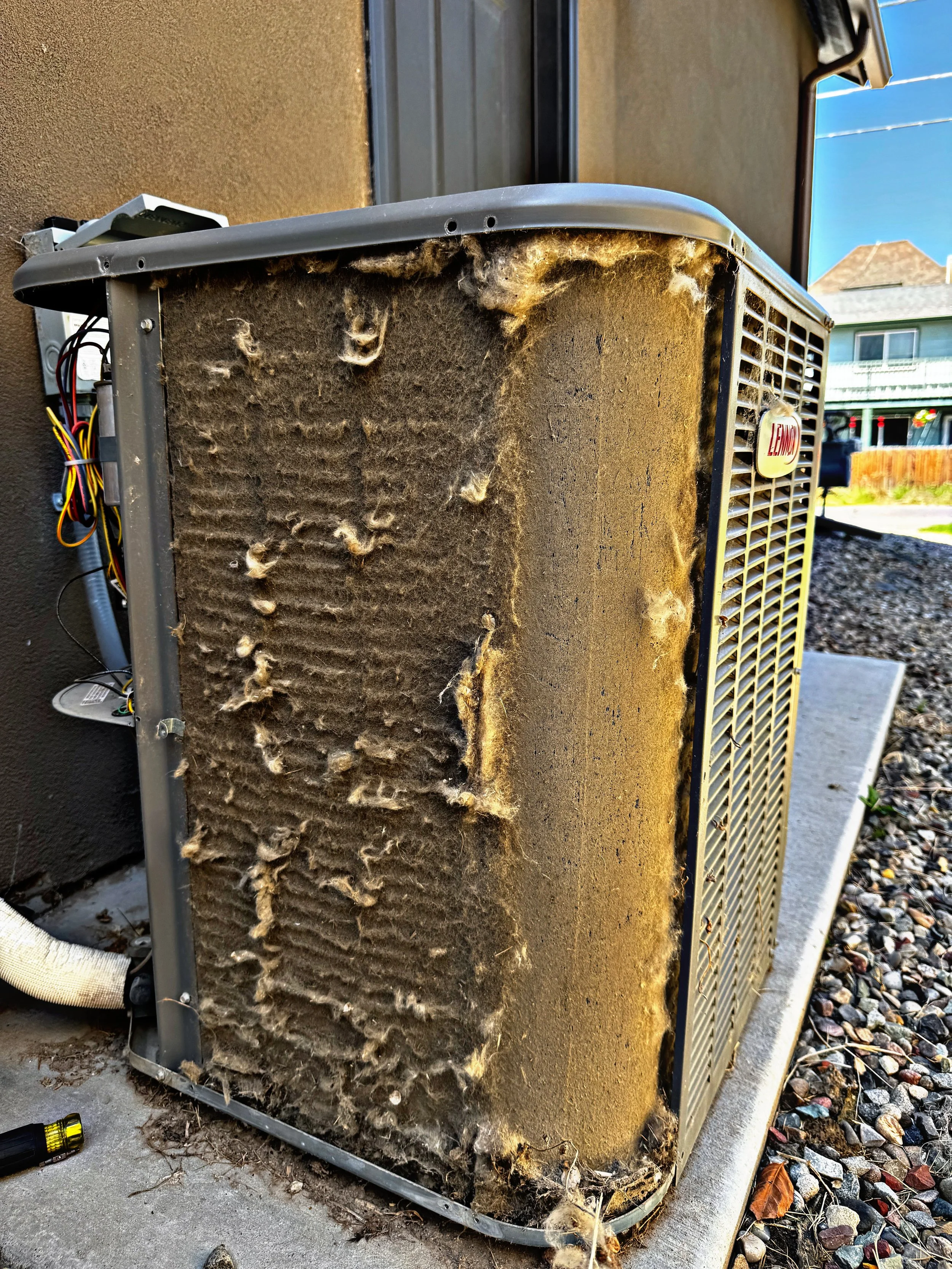 Dirty and neglected outdoor air conditioning unit covered in dirt, debris, and dust with exposed wires on the side, positioned outside a building next to gravel and concrete ground.