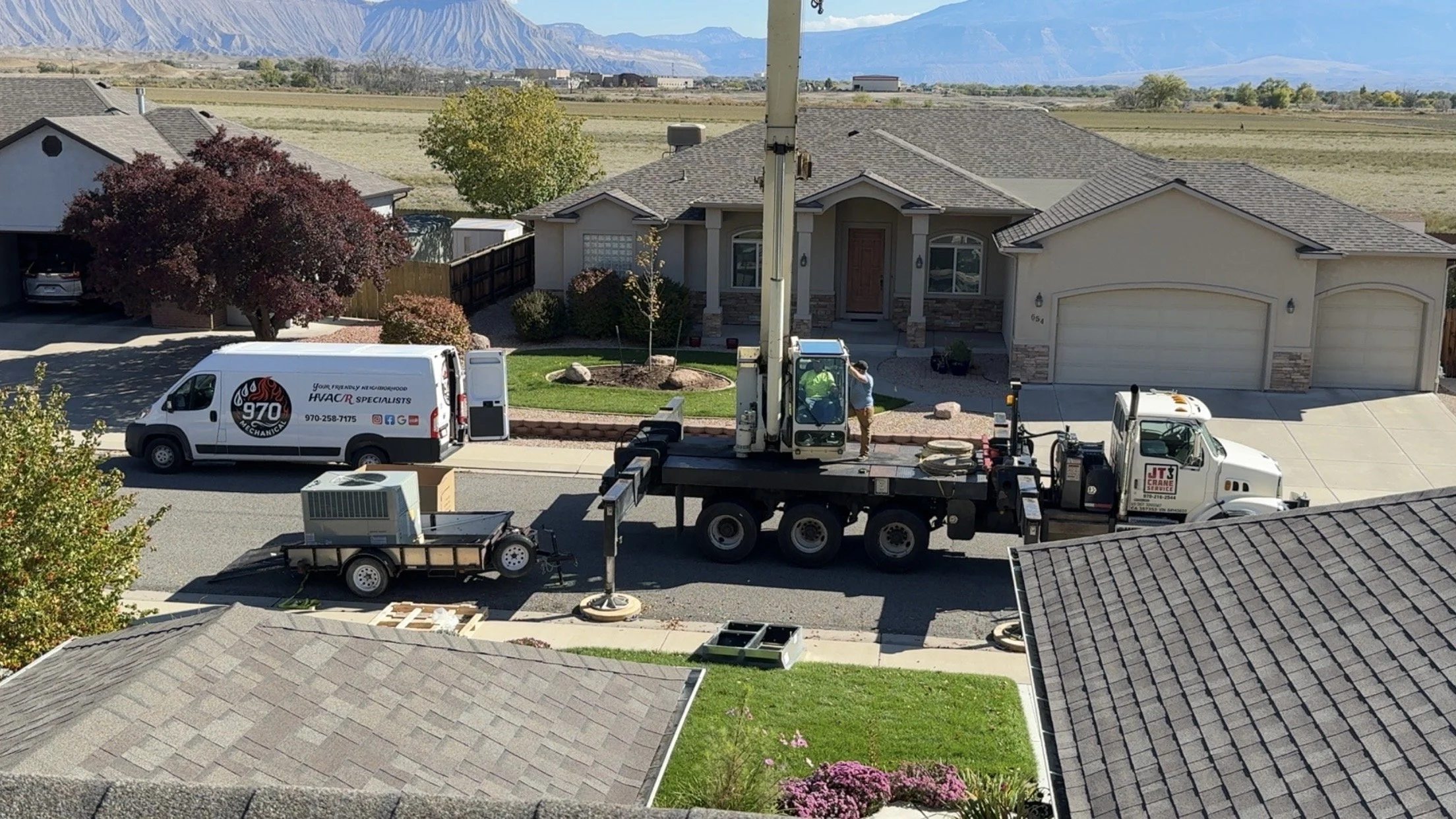 A residential street with a house having a landscaped front yard, trees, and mountains in the background. A crane truck and a service van are parked on the street, with a worker standing near the crane.
