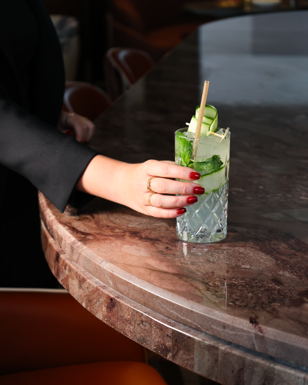 A person with red nail polish and gold rings holds a glass of cucumber cocktail on a marble bar counter. The drink contains cucumber slices and a green garnish with a wooden stir stick.
