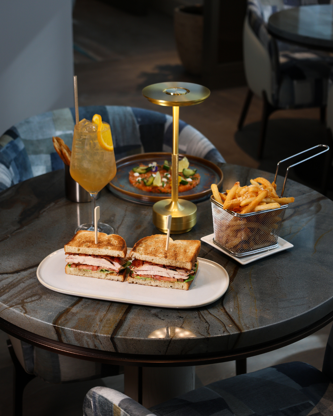 A meal on a dark marble table features a sandwich cut in half with turkey, lettuce, and tomato, a French fry basket, a colorful pizza with vegetables, and a yellow cocktail with ice and lemon slices.