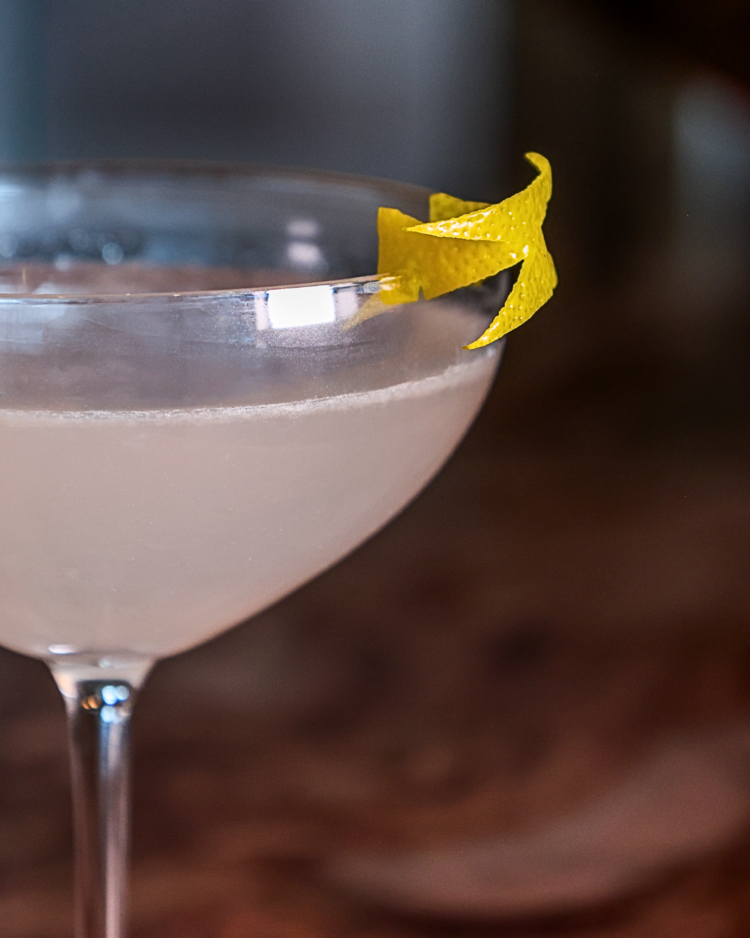 A close-up of a cocktail with a lemon peel garnish in a coupe glass. The drink appears to be light pink or pale purple.