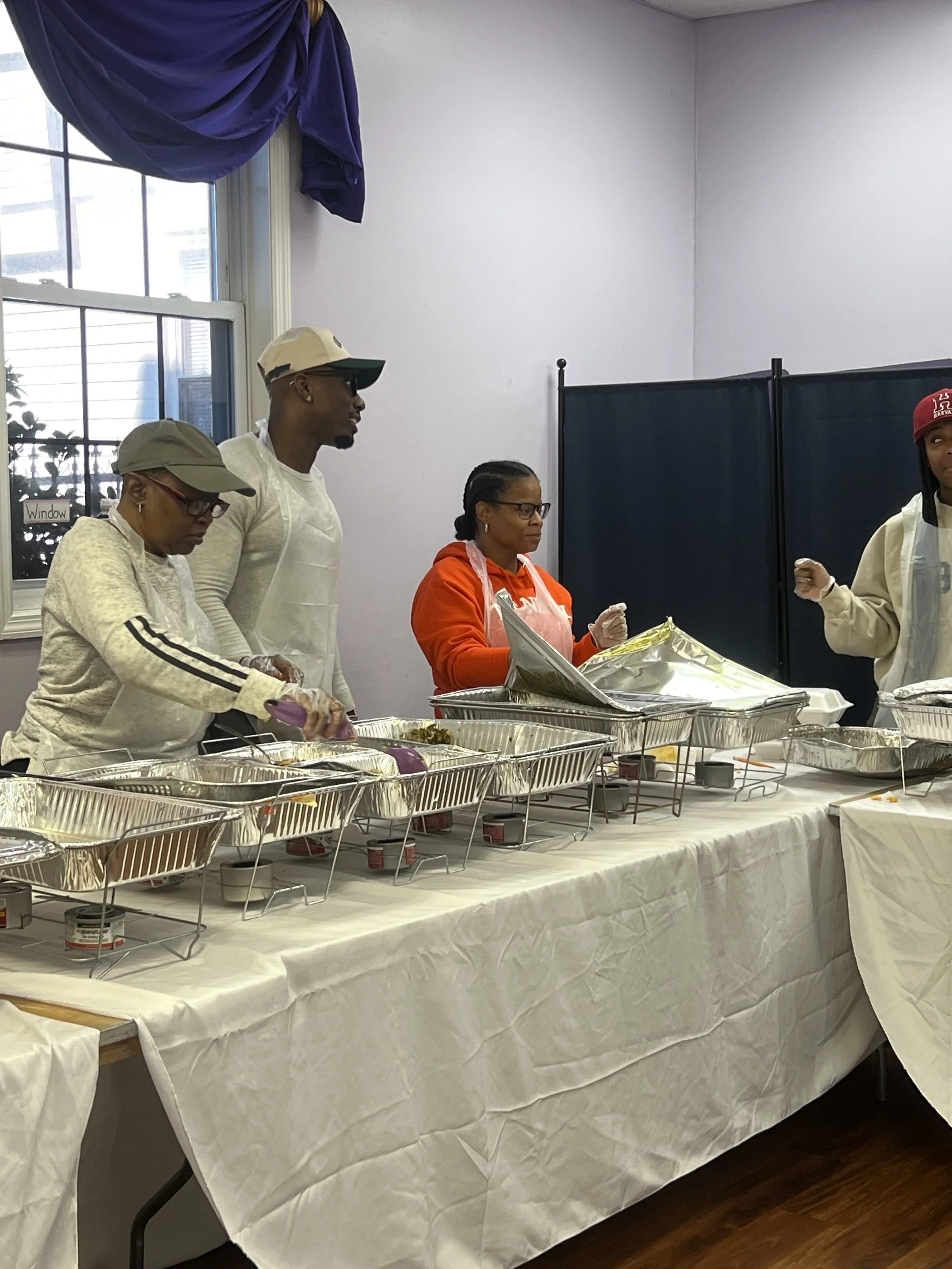 People serving food at a buffet table inside a room with a window, blue curtains, and a purple curtain rod.
