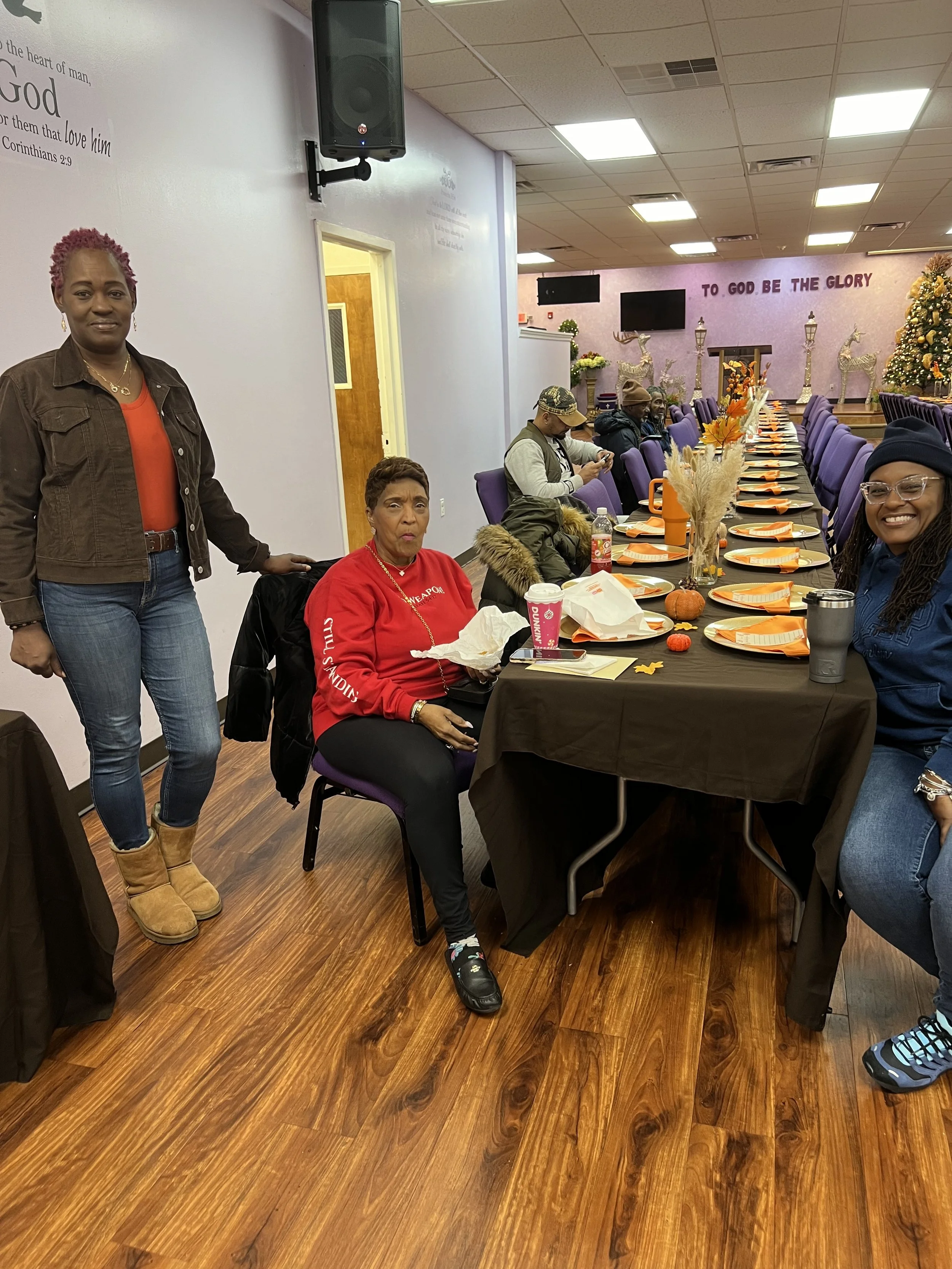 A group of people sitting and standing around a long decorated table in a church or community room, with a Christmas tree and festive decorations in the background.
