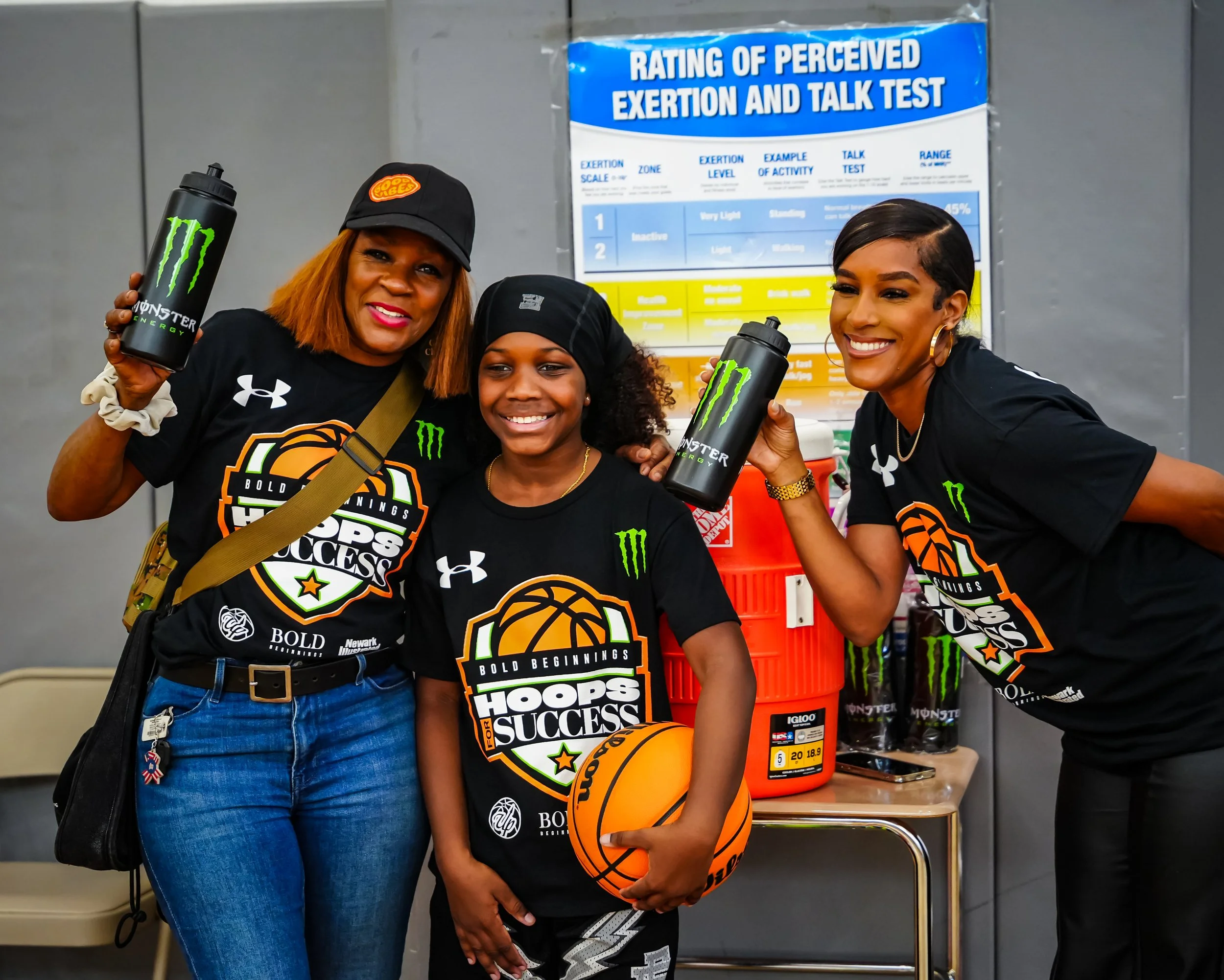 Three women and a girl smiling, holding Monster energy drink bottles, wearing matching black t-shirts with basketball-themed design and text, inside a gym or sports facility with a poster about exertion testing in the background.