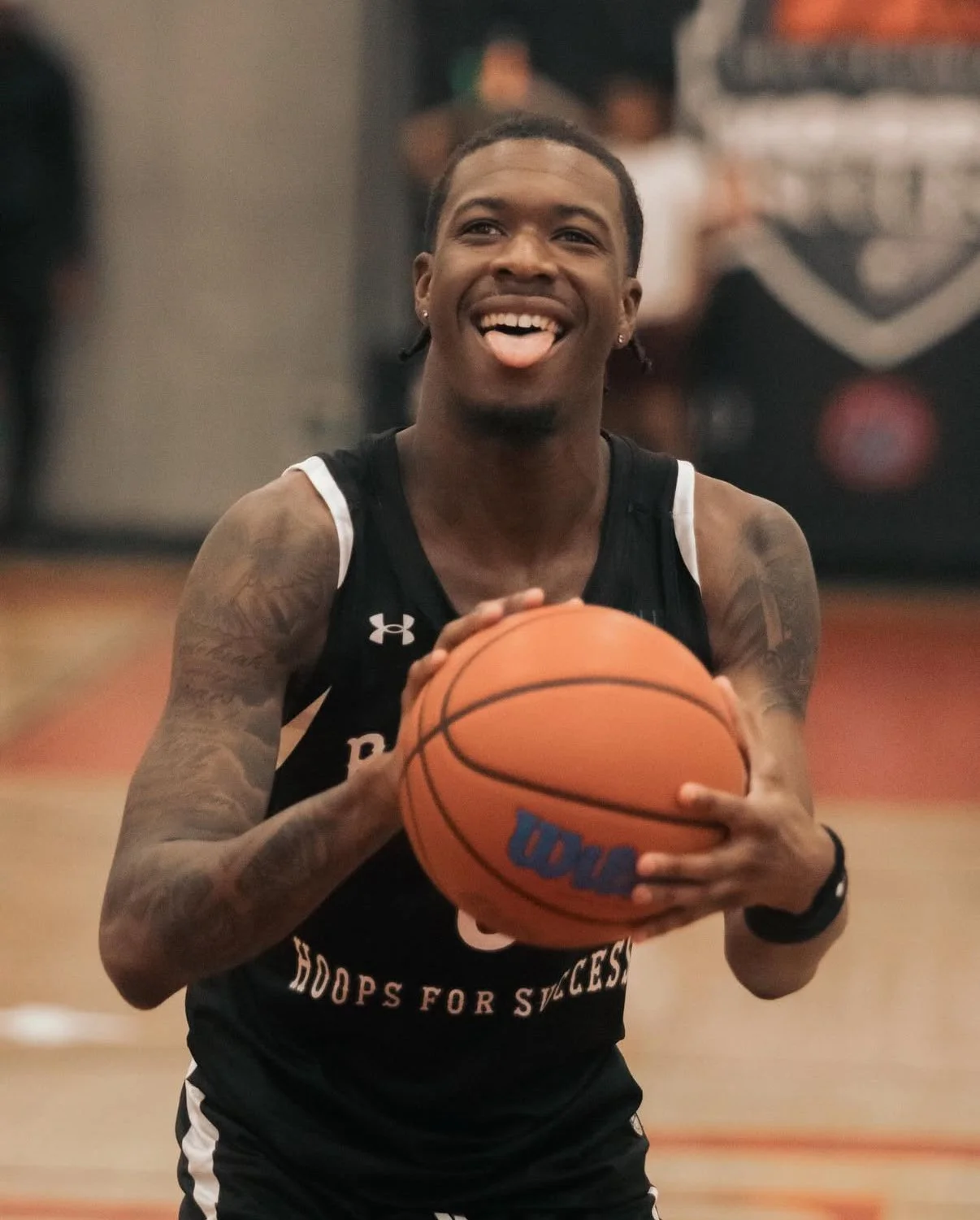 A man smiling and sticking out his tongue while holding a basketball on an indoor court.