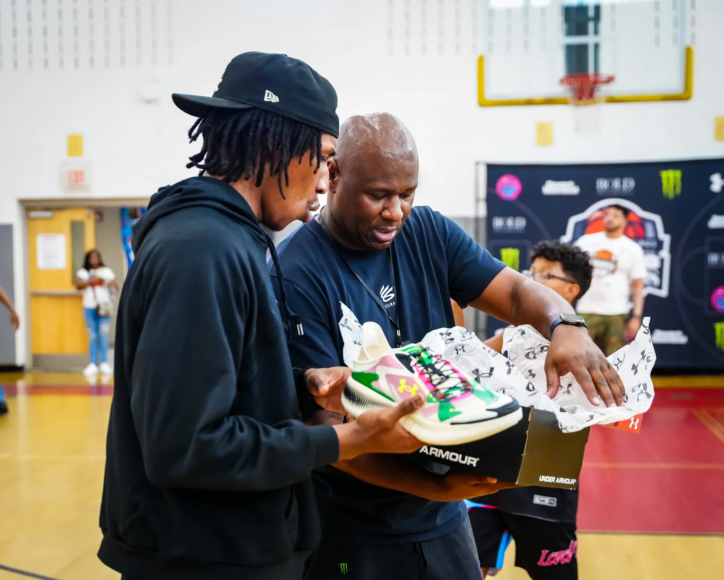 Two men are holding a colorful pair of Under Armour sneakers, examining them inside a gymnasium with a basketball hoop and a backdrop with logos in the background.