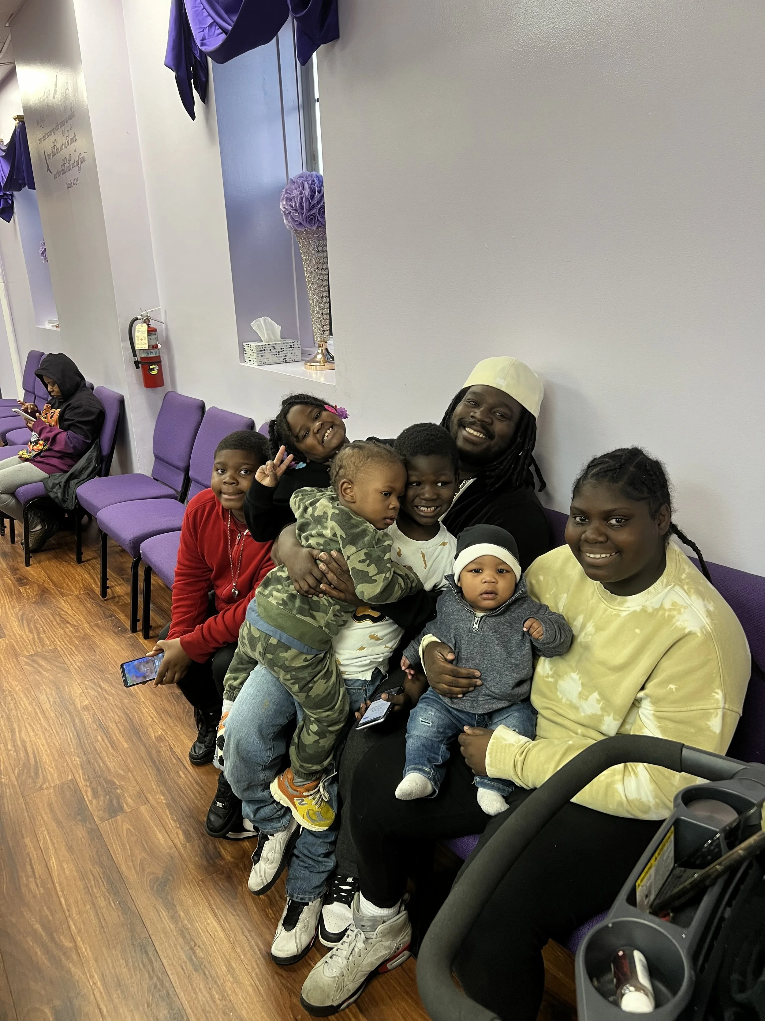 A man with dreadlocks, wearing a white hat, is sitting on a purple chair holding two children. Four other children, some making peace signs, are sitting or standing close by, smiling and posing for the photo. Inside a room with purple chairs along th