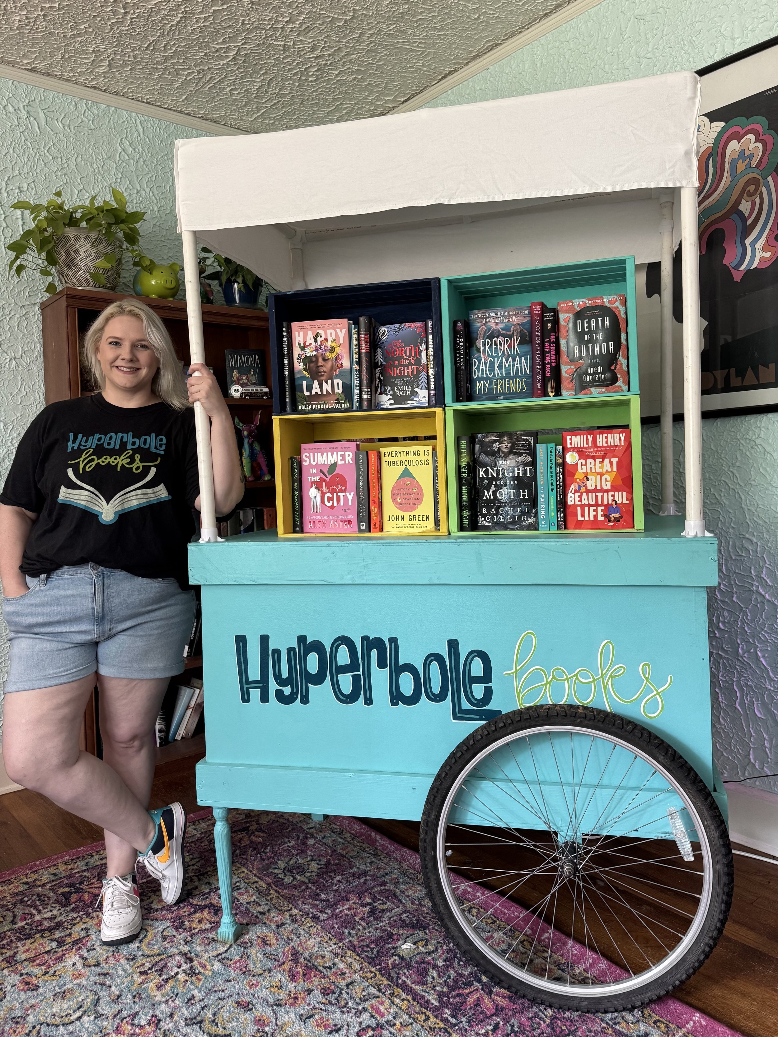 a smiling girl stands next to a turquoise cart with a white canopy that reads "Hyperbole Books." On the cart are colorful crates displaying fiction and nonfiction books.