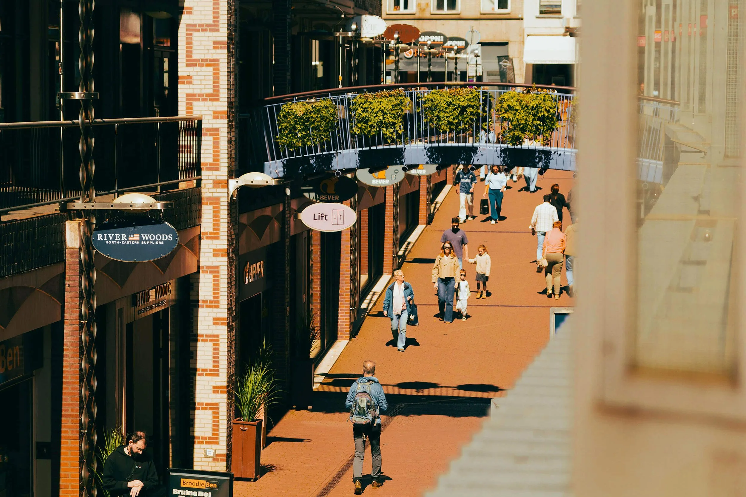 A busy infill development city street with pedestrians walking along a sidewalk lined with shops and businesses. The scene includes a balcony with potted plants and signage for various stores and restaurants.