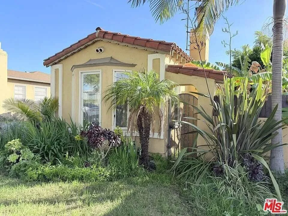 Yellow house with red tile roof, front yard with lush green tropical plants, palm trees, and bushes, bright blue sky in the background.