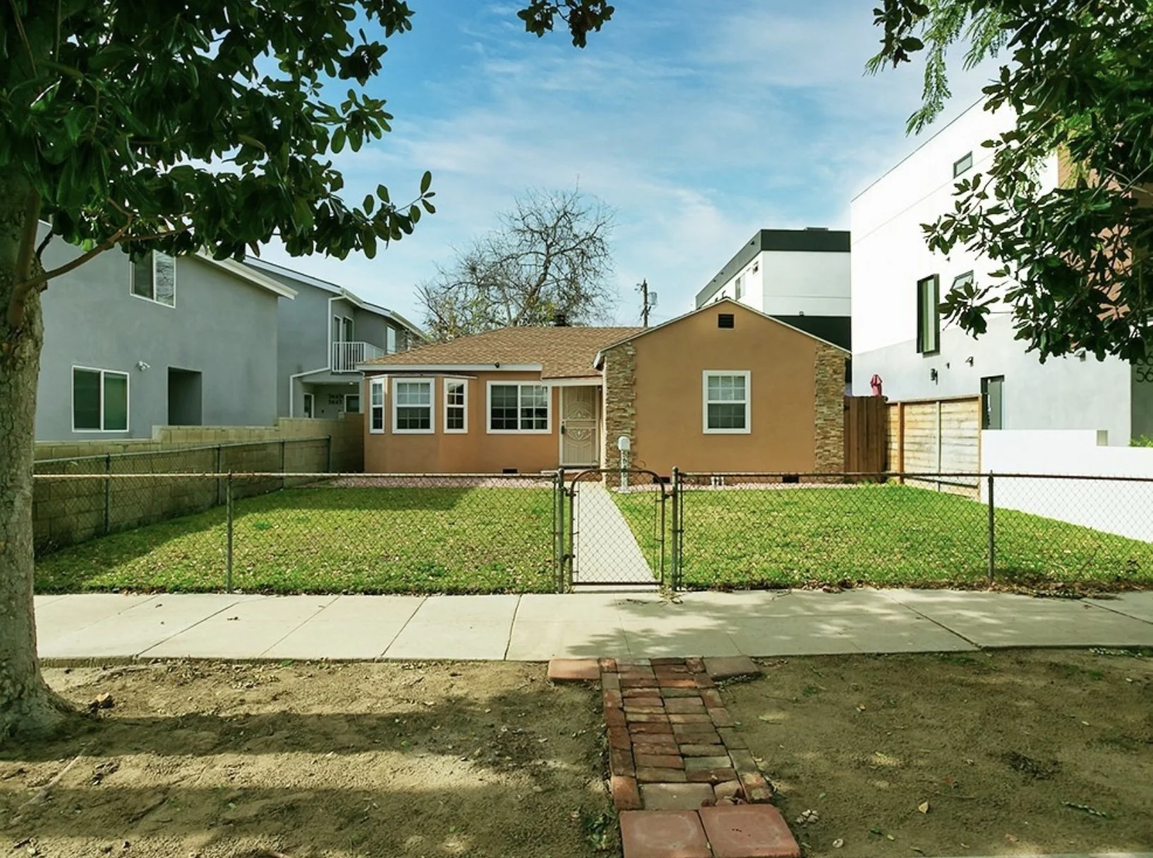 A small front yard with a chain-link fence and gate, grass, a concrete sidewalk, and brick pathway. In the background, there are two houses, one beige with stone accents and one grey apartment-like building. There are trees and a blue sky with some c