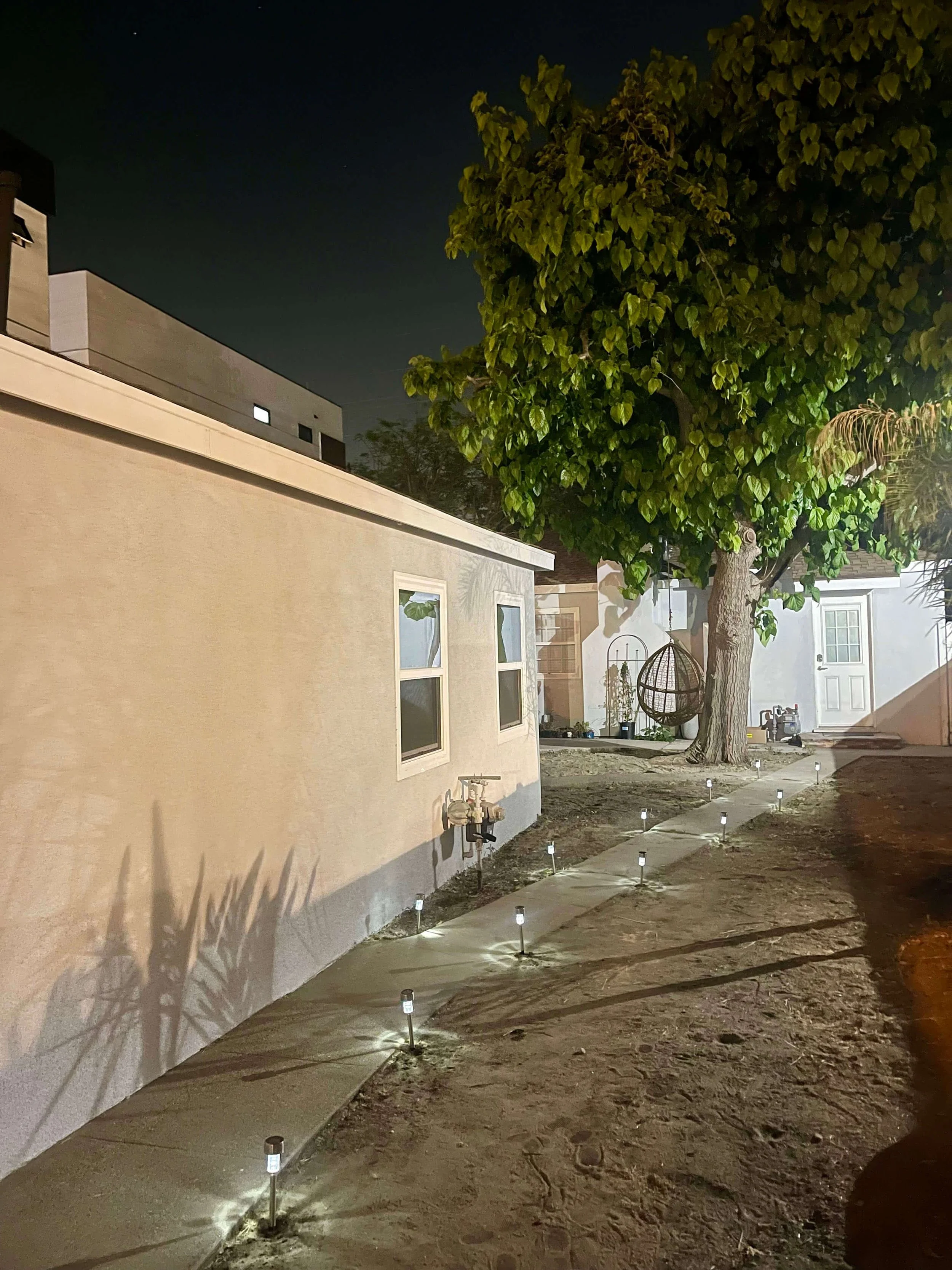 Nighttime exterior view of residential backyard with a large leafy tree, a beige house with white-framed windows, pathway lights along a concrete walkway, and a hanging wicker chair, all illuminated by ground lights.