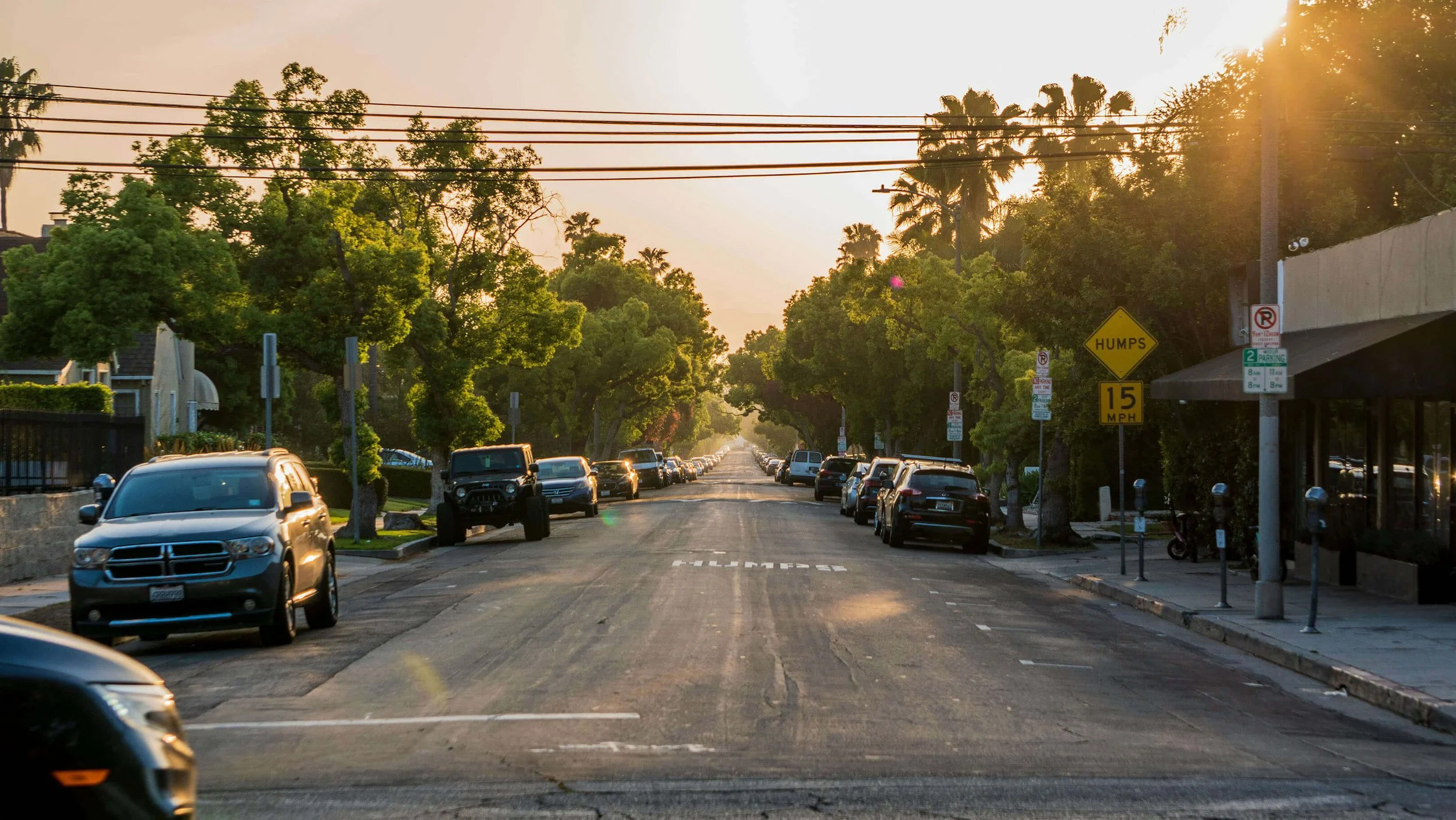 A city street during sunset with parked cars on both sides, trees lining the street, and signs indicating a humps and parking regulations.