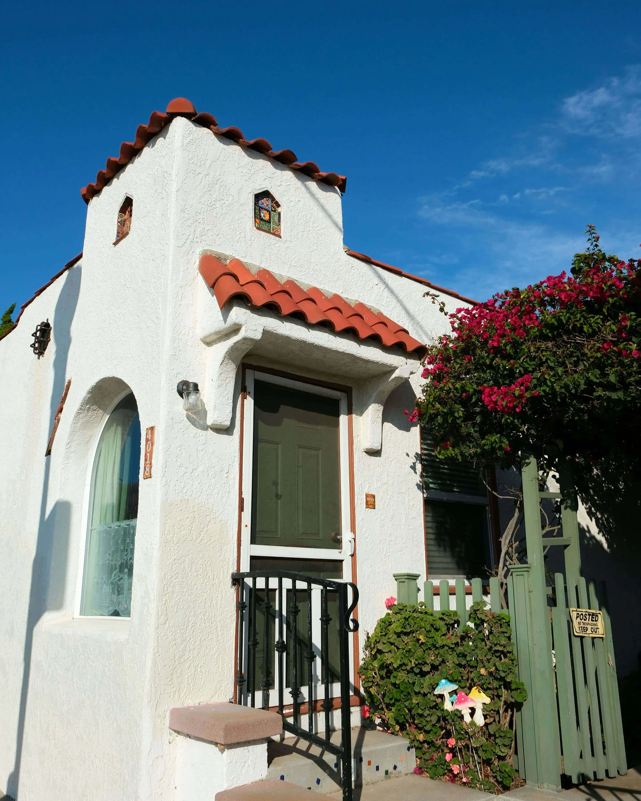 A white stucco house with a red tile roof, small stained glass windows, and a green door. There is a small front porch with a black railing, potted plants, and colorful mushroom garden decorations. A green picket fence and pink flowering bushes are visible beside the house, with a sign that says 'No Trespassing Keep Out'.