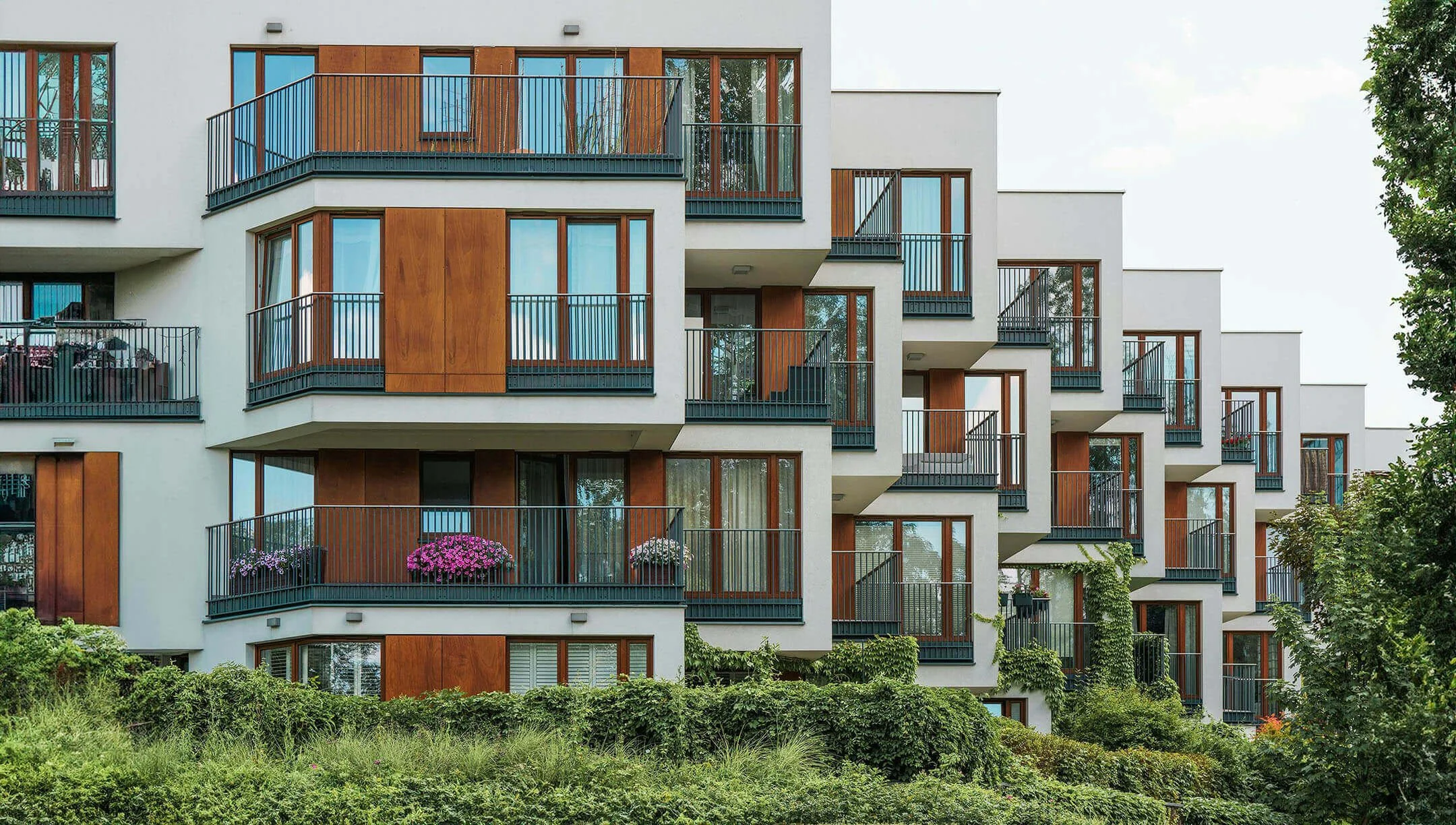 A modern multi-story apartment building with white and wood exterior walls, multiple balconies with black railings, and large glass windows. There are some potted plants and flowers on the balconies, and trees with green foliage surround the building. The sky is clear or lightly cloudy.