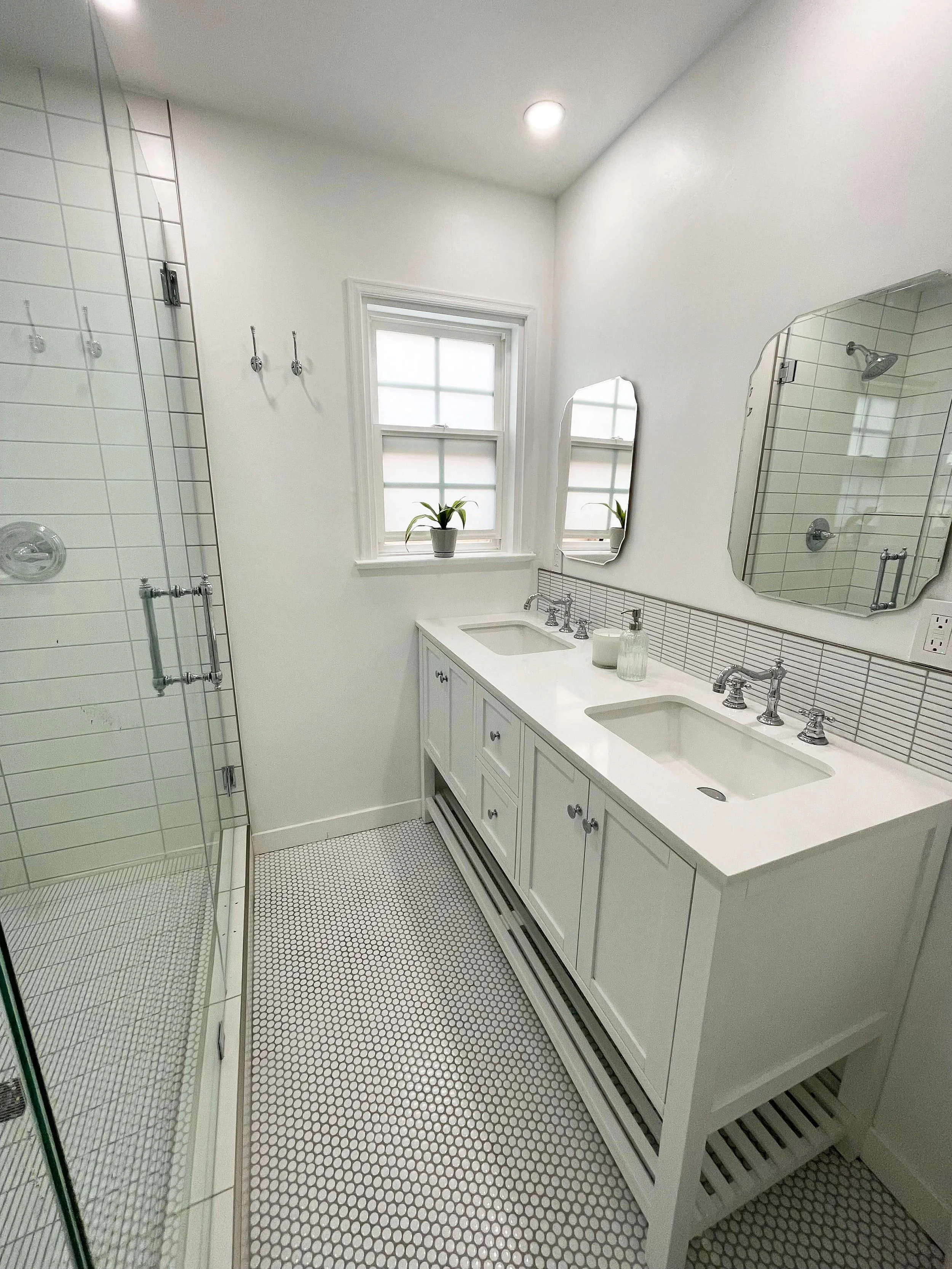 Clean bathroom with a white vanity, two cabinets, two sinks, mirrored medicine cabinet, and a tiled shower with glass door and white tiles, two windows with potted plants, and a ceiling light.